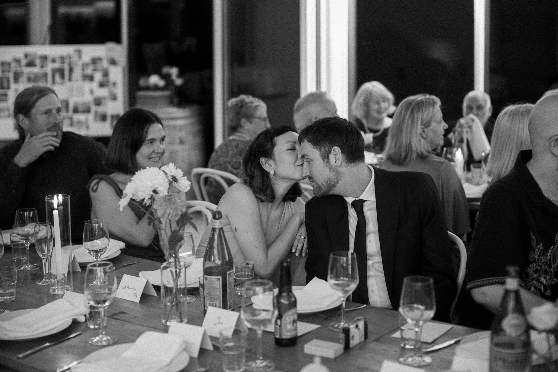 A black-and-white photo of a wedding reception with guests seated at tables, including a woman kissing a man on the forehead, surrounded by glasses, flowers, and table cards.