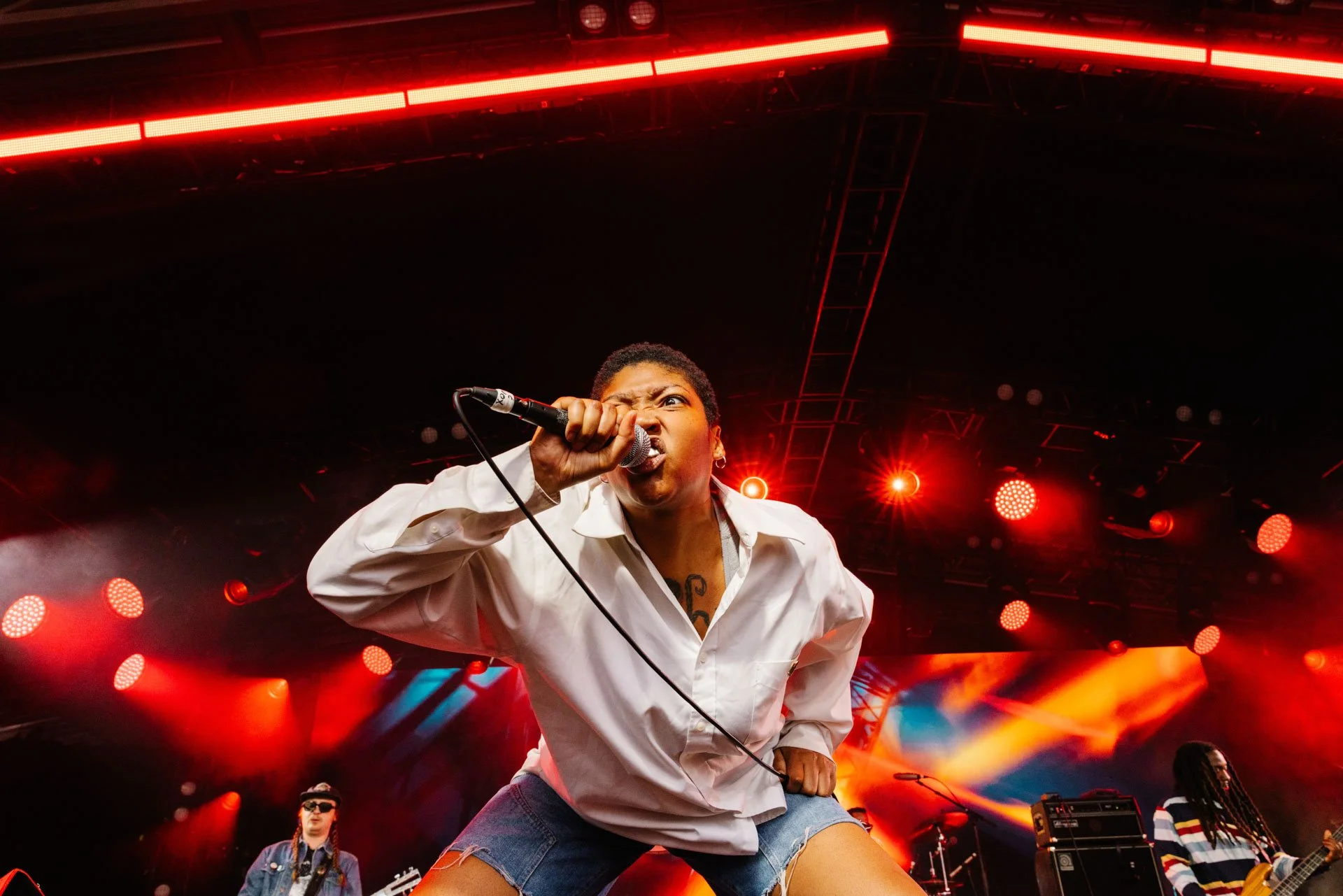 A female singer performing passionately on stage with red stage lighting, wearing a white shirt and shorts, holding a microphone close to her mouth, with a band playing in the background.