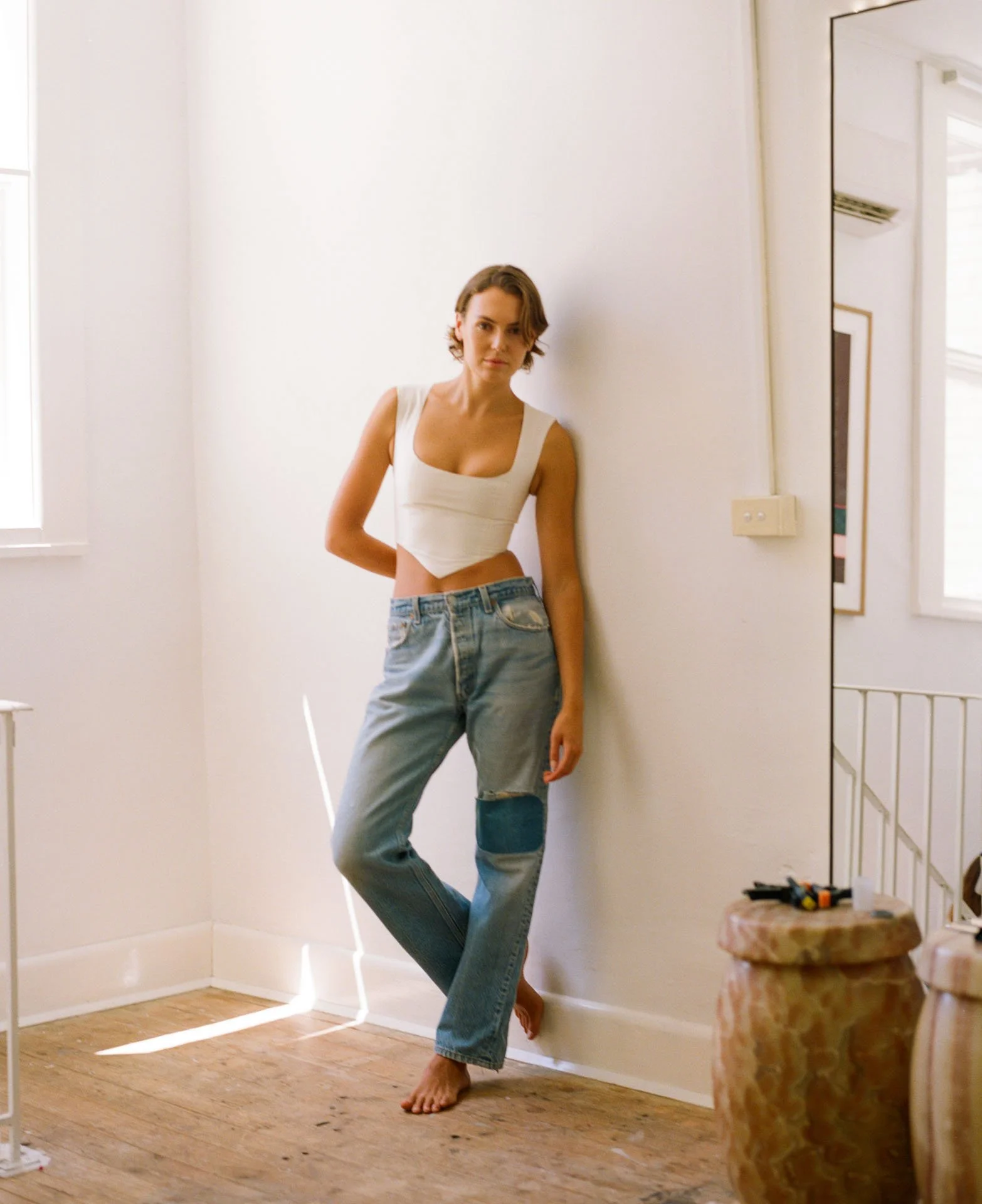 A woman with short hair wearing a white crop top and high-waisted jeans standing barefoot in a bright room with wooden flooring.