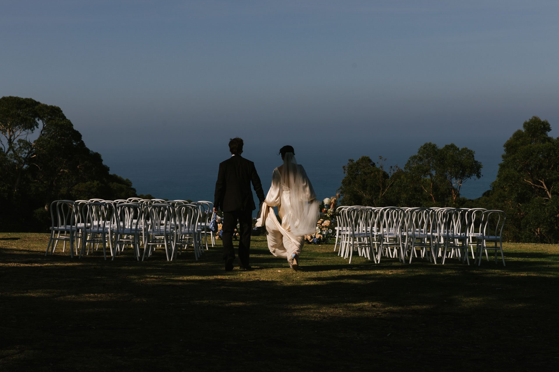 A bride and groom walking hand in hand down an outdoor aisle at sunset, with empty chairs on either side and trees in the background.