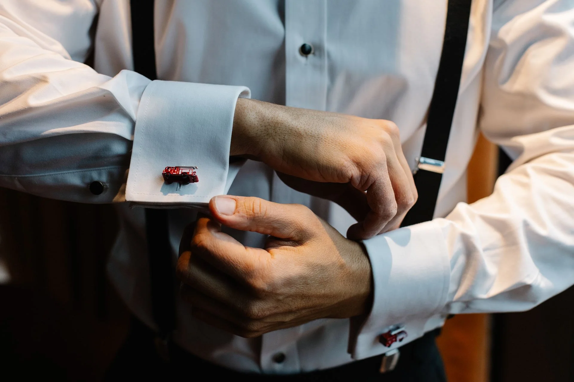 A person in a white dress shirt with French cuffs is attaching a red fire truck lapel pin to their cuff, with another person assisting. The person is also wearing black suspenders.