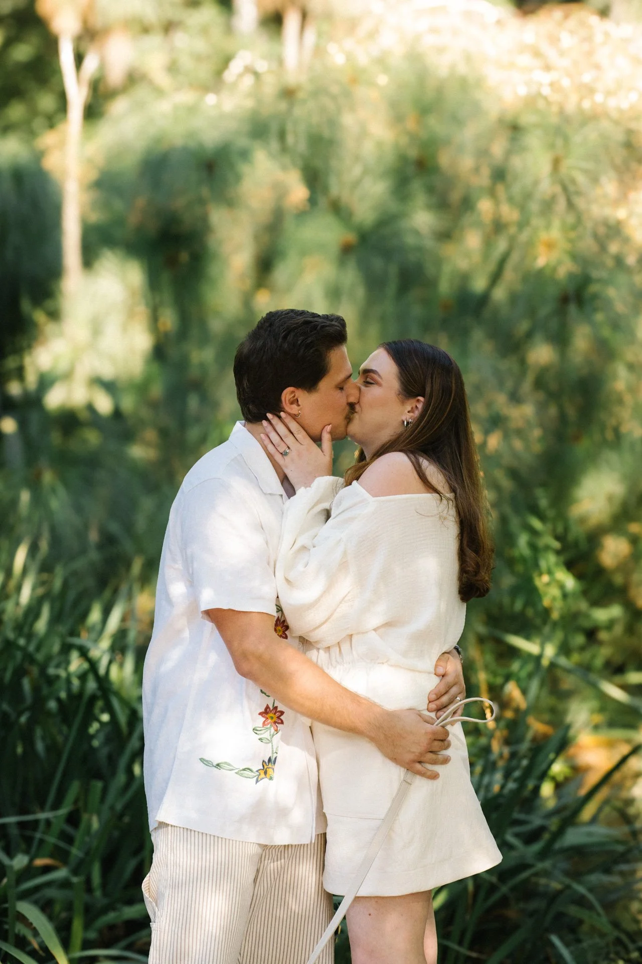 A couple sharing a kiss outdoors surrounded by lush greenery.