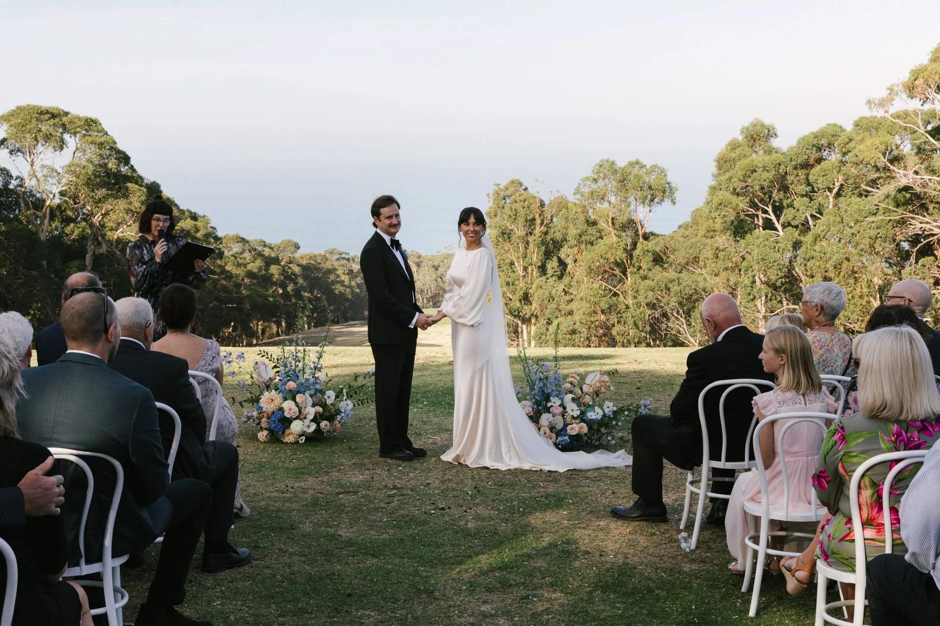 A wedding ceremony outdoors with a bride and groom holding hands and smiling, surrounded by seated guests, with a woman officiant reading from a book and a scenic background of trees and open sky.