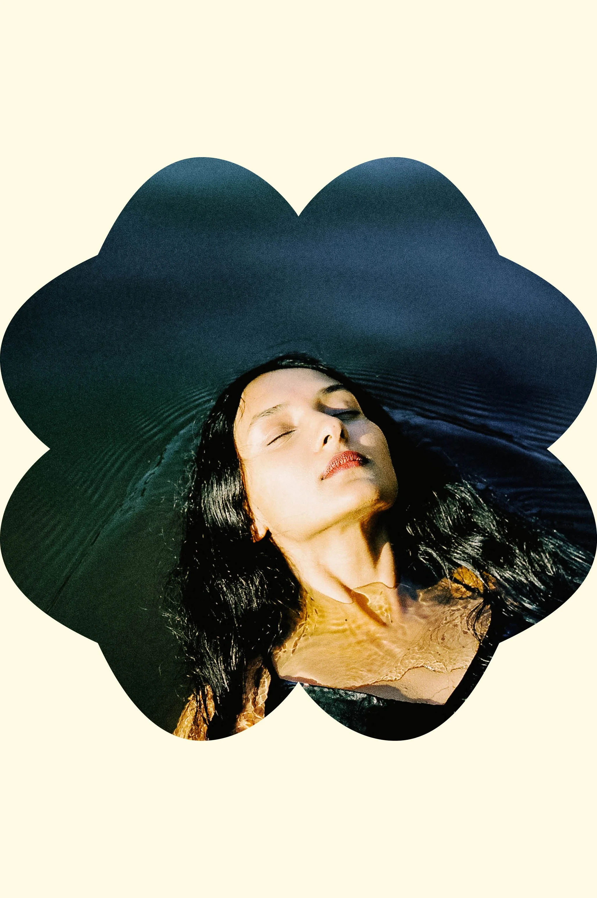 A woman with black, wet hair lying on a large, wide-brimmed black hat, with her eyes closed and her face relaxed.