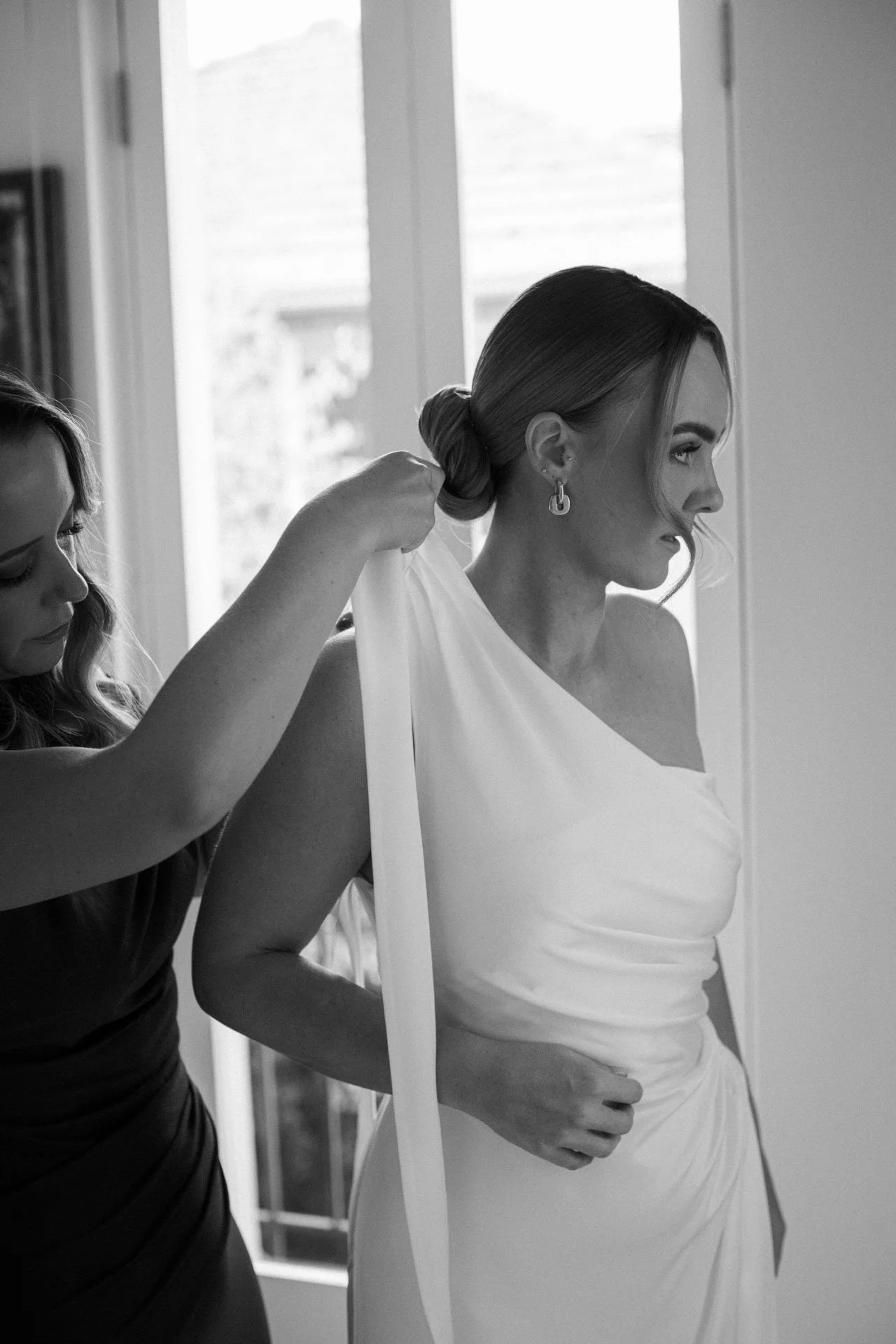 A woman in a white dress is getting ready, with another woman assisting her with her hair, in a room with large windows.