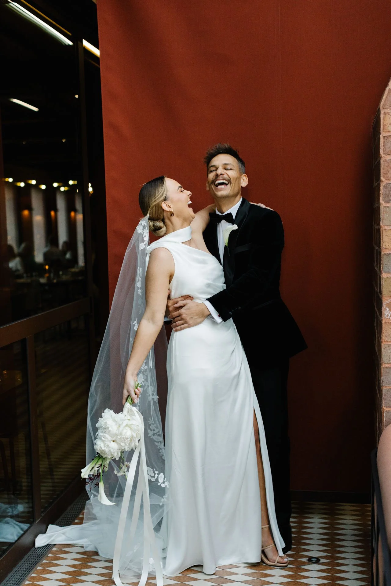 A bride and groom sharing a joyful moment indoors, dressed in wedding attire, with the bride holding a bouquet of white flowers and the groom smiling.