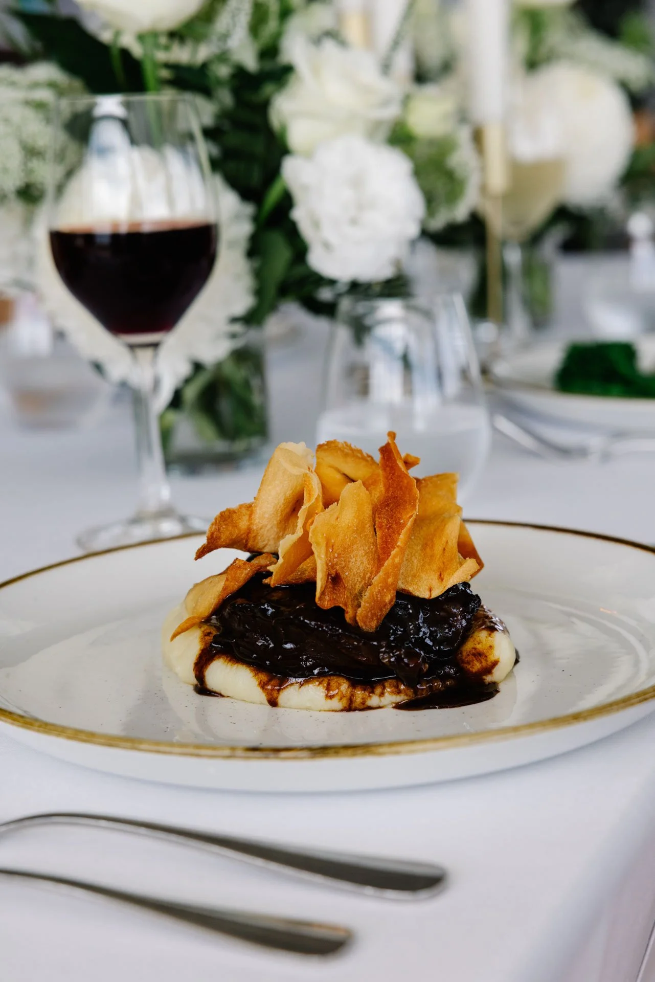 Dessert with a dark fruit compote, gallette crust, and garnished with crispy thin slices of sweet potato, served on a white plate with gold trim.