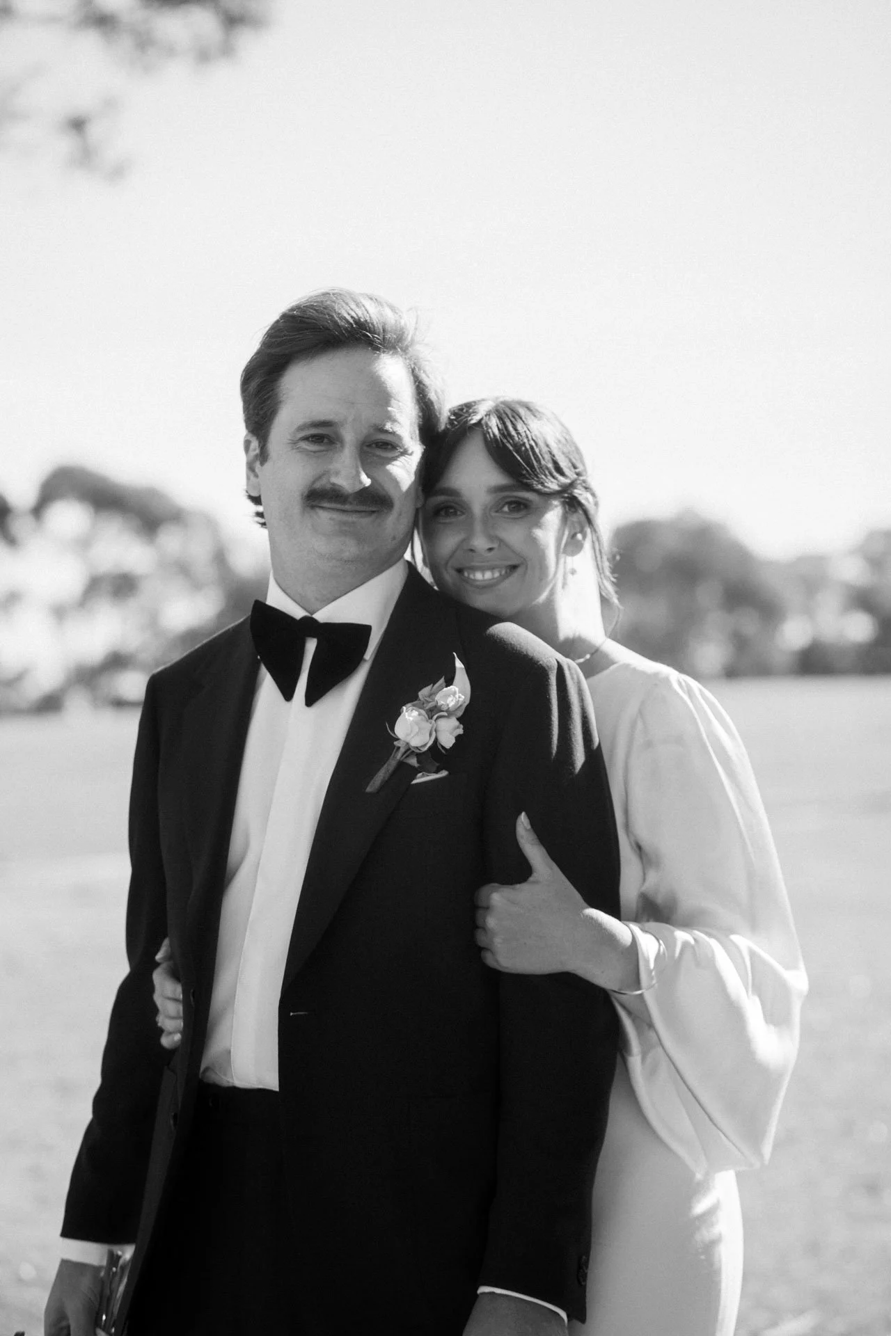 A black and white photo of a groom and bride smiling and hugging outdoors, with a blurred background of trees and open sky, dressed in wedding attire.