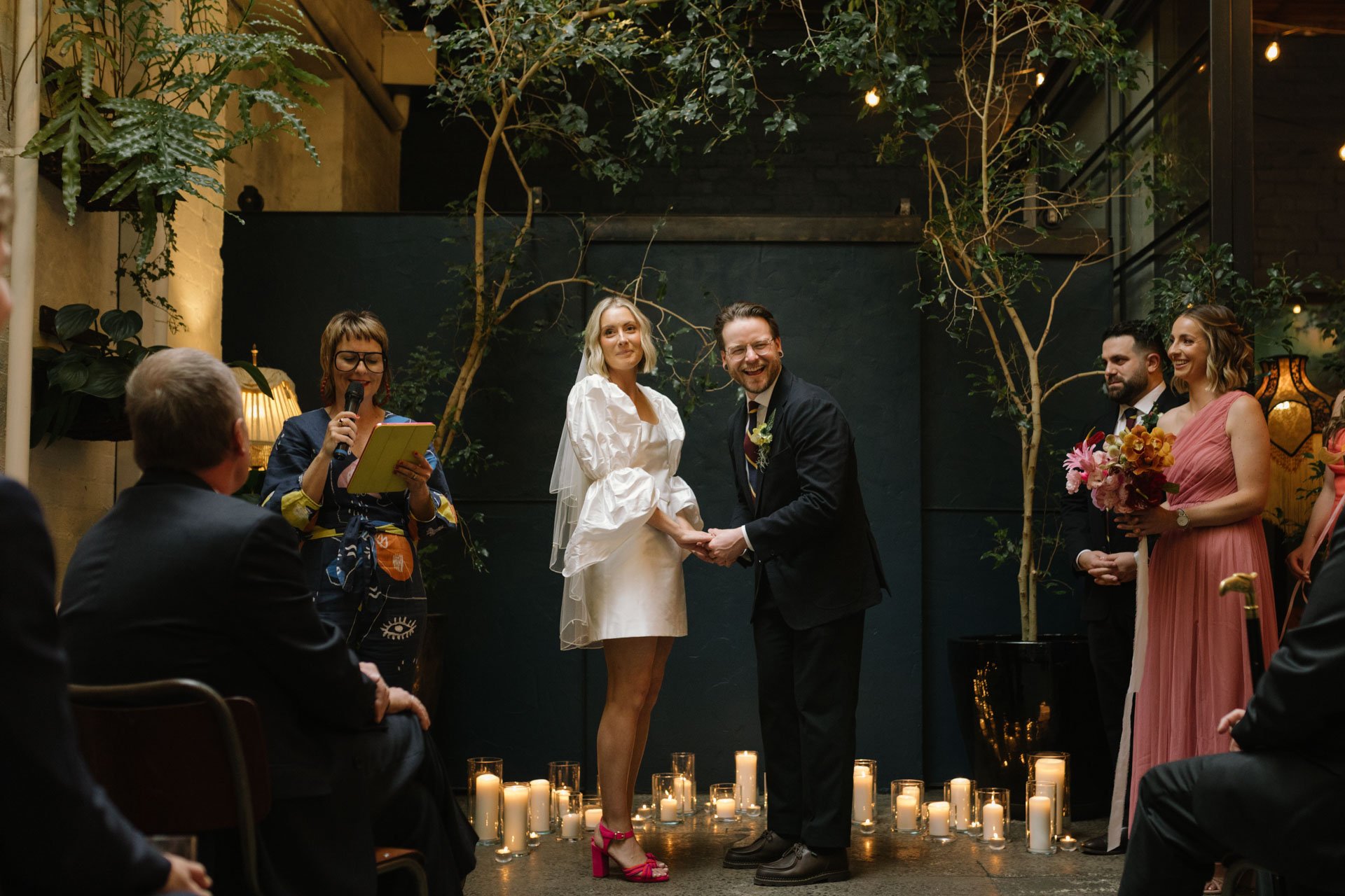 A wedding ceremony with a bride and groom holding hands, facing each other, surrounded by several guests. The bride is wearing a white dress with puffy sleeves and pink heels, and the groom is dressed in a black suit with a boutonniere. A officiant w