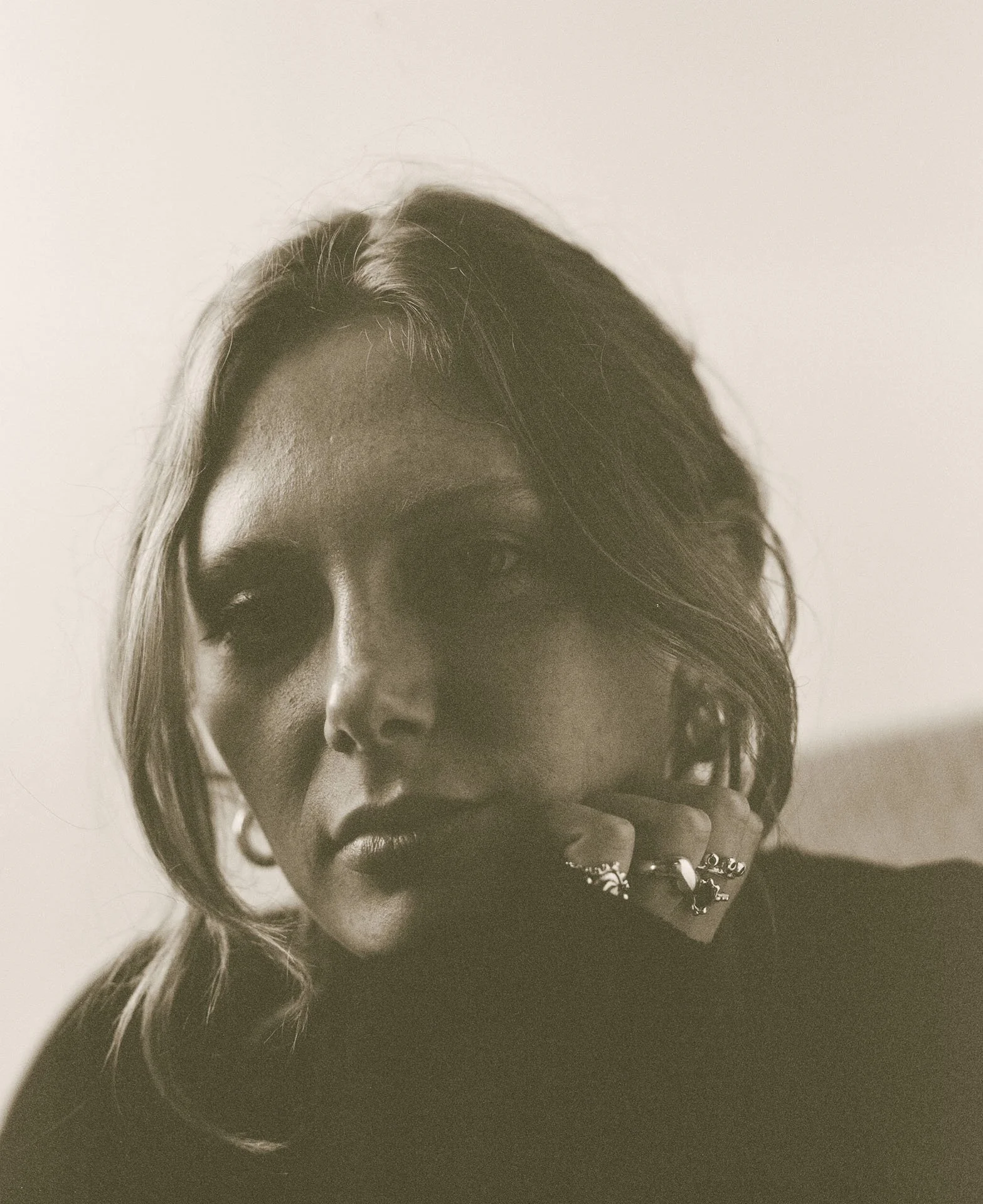 A black and white photo of a woman with wavy hair, dark lipstick, and multiple rings on her fingers, touching her lips.