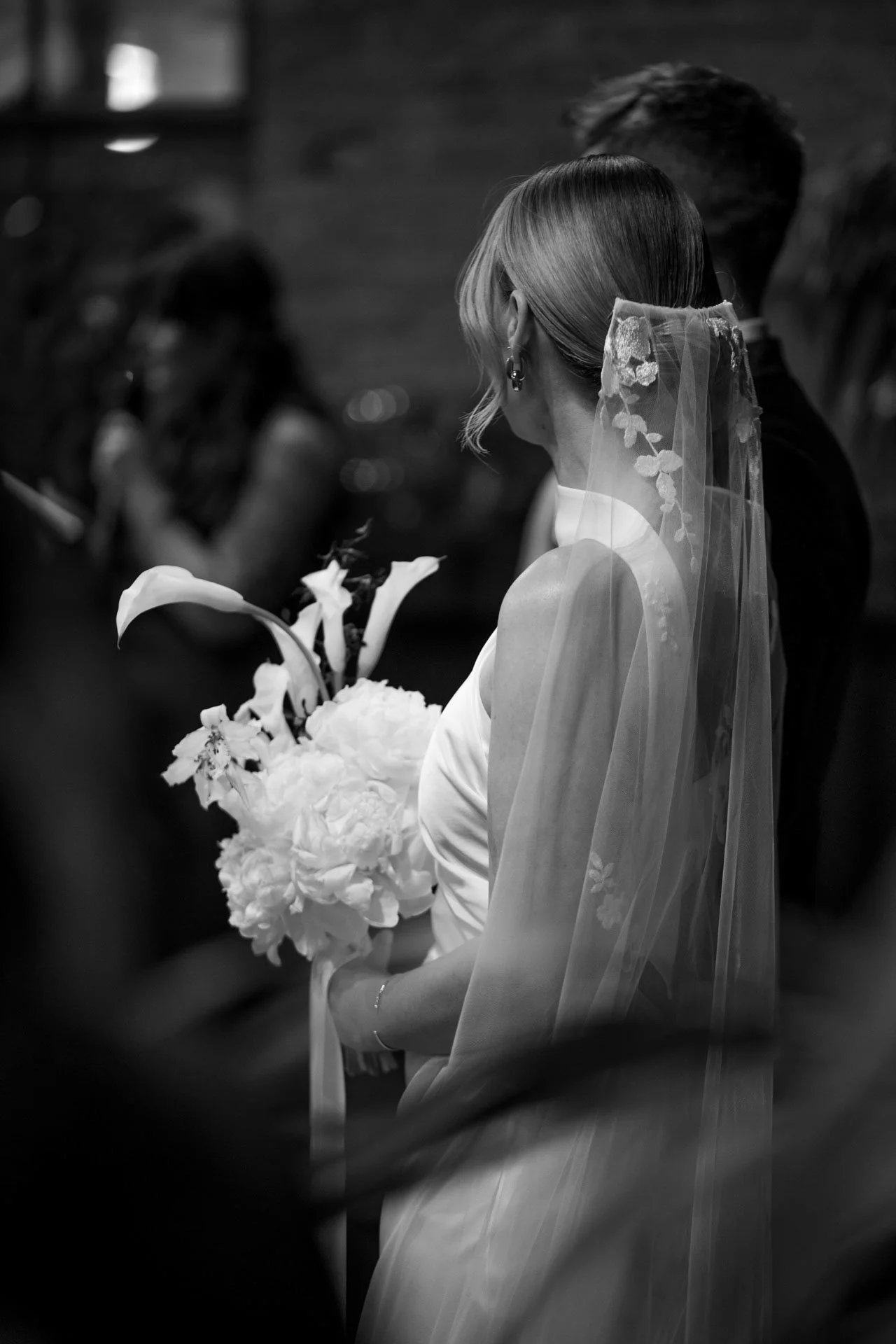 A bride holding a bouquet of flowers, wearing a wedding dress and veil, facing a groom in a black suit during a wedding ceremony.