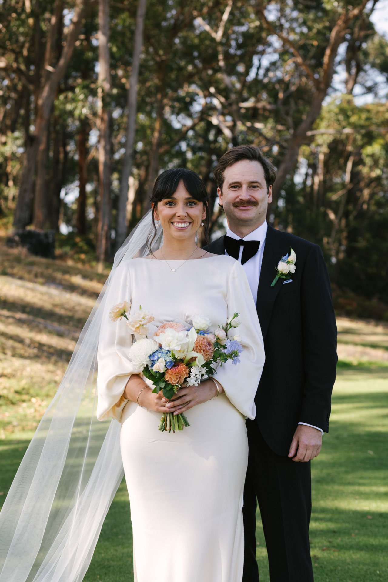 A bride and groom standing outdoors in a park, smiling, with trees in the background. The bride wears a white dress and veil, holding a colorful bouquet of flowers. The groom wears a black tuxedo with a white shirt and black bow tie, with a boutonnie