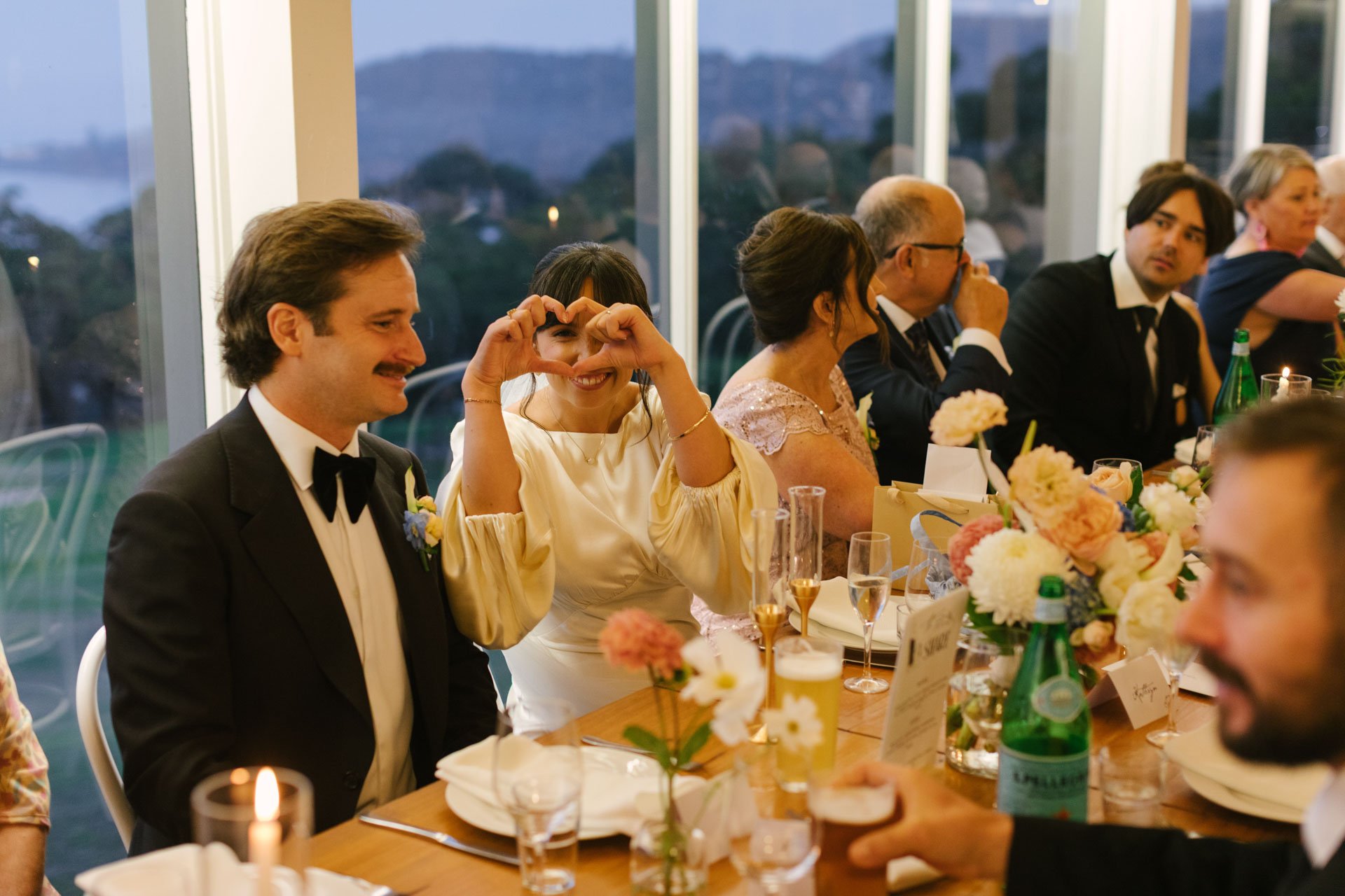 People attending a formal dinner party, sitting at a long table decorated with flowers and candles, with mountains and a lake visible outside the windows in the background.