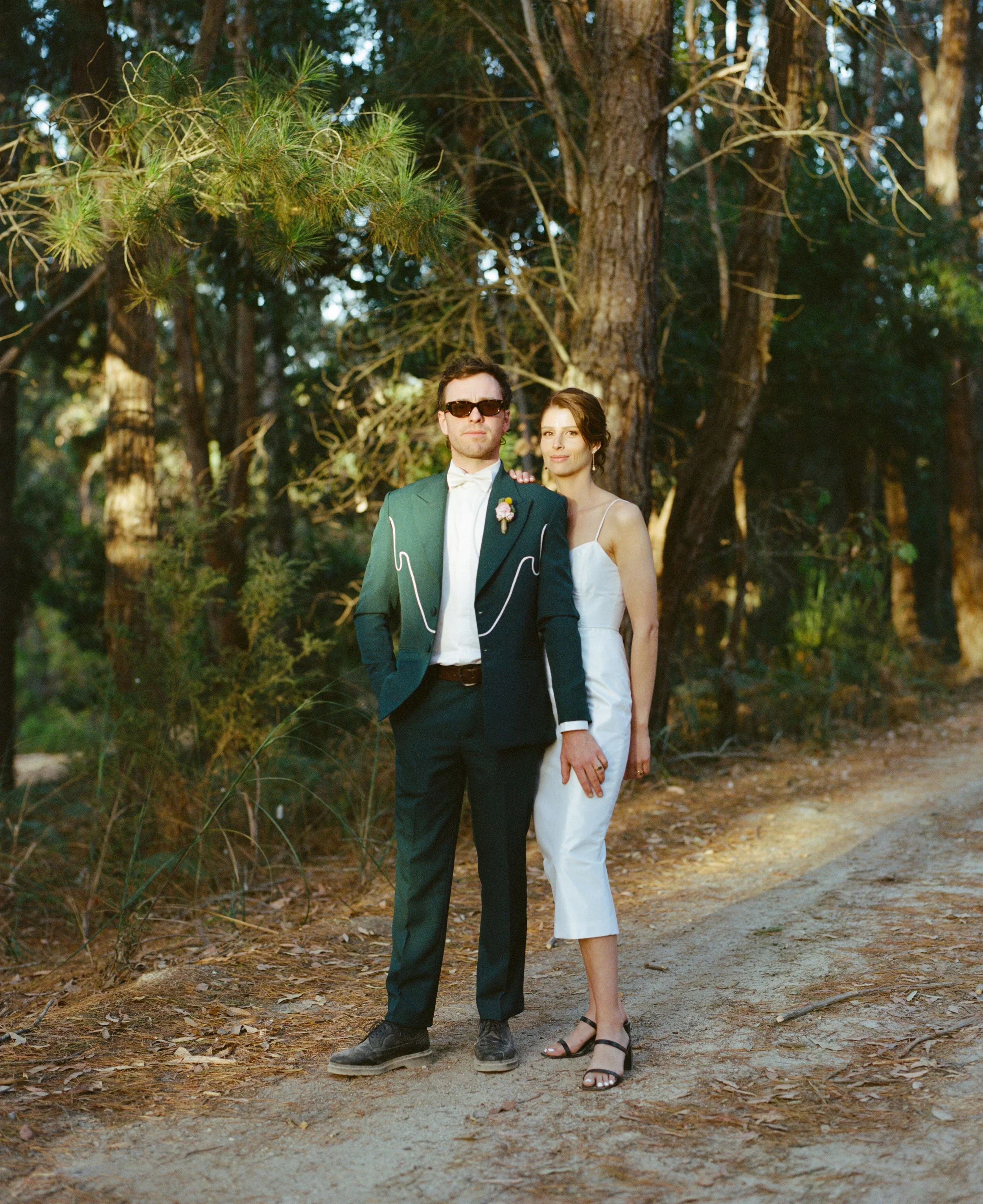 A man in a tuxedo and a woman in a white dress standing together outdoors in a forested area.