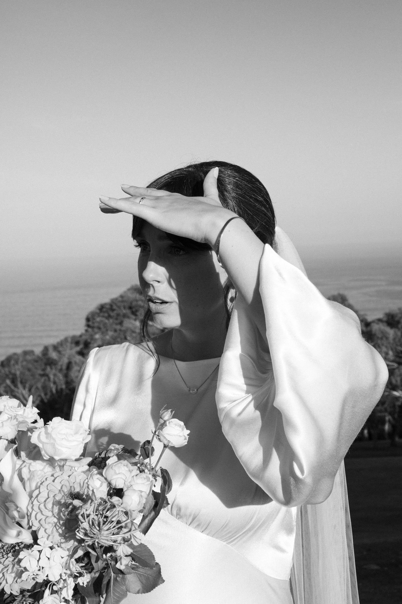 A woman in a white dress holding a bouquet of flowers outdoors, shielding her eyes from the sun with her hand.