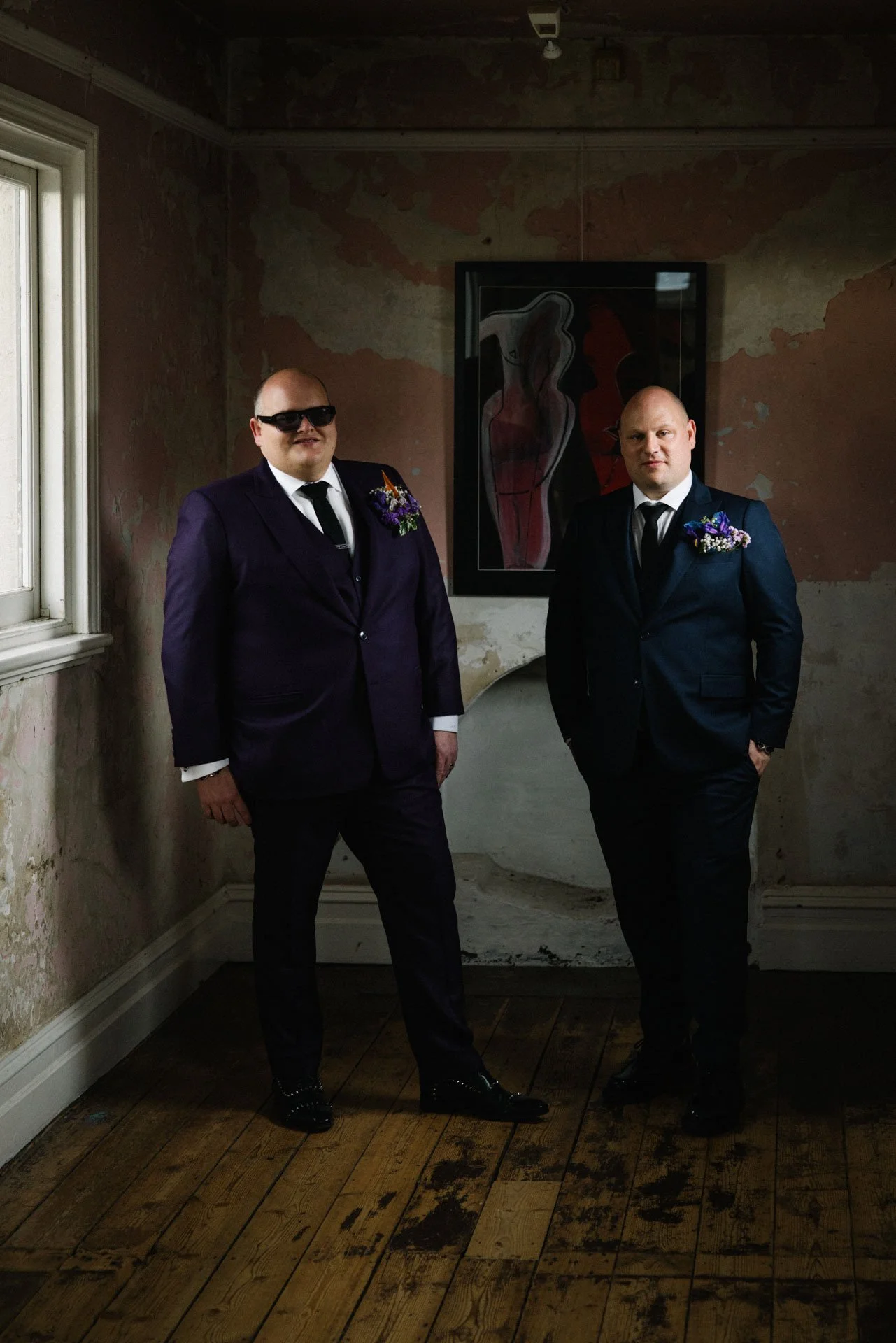 Two men in suits standing in a room with peeling paint walls and a wooden floor, one wearing sunglasses and both with flowers on their lapels.