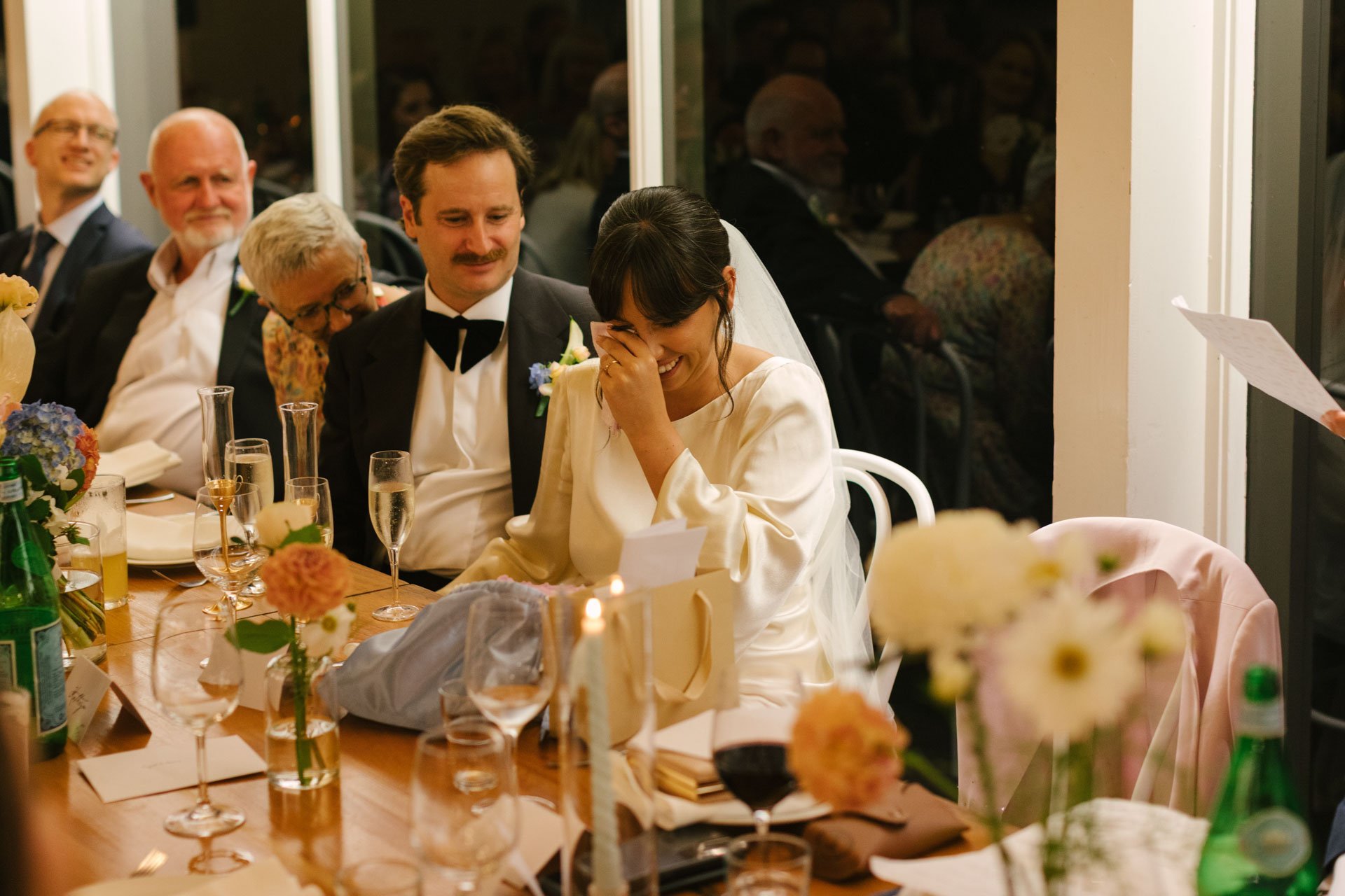 A bride and groom sitting at a table during a wedding reception, with the bride laughing and covering her face, surrounded by guests and decorated tables with flowers and drinks.