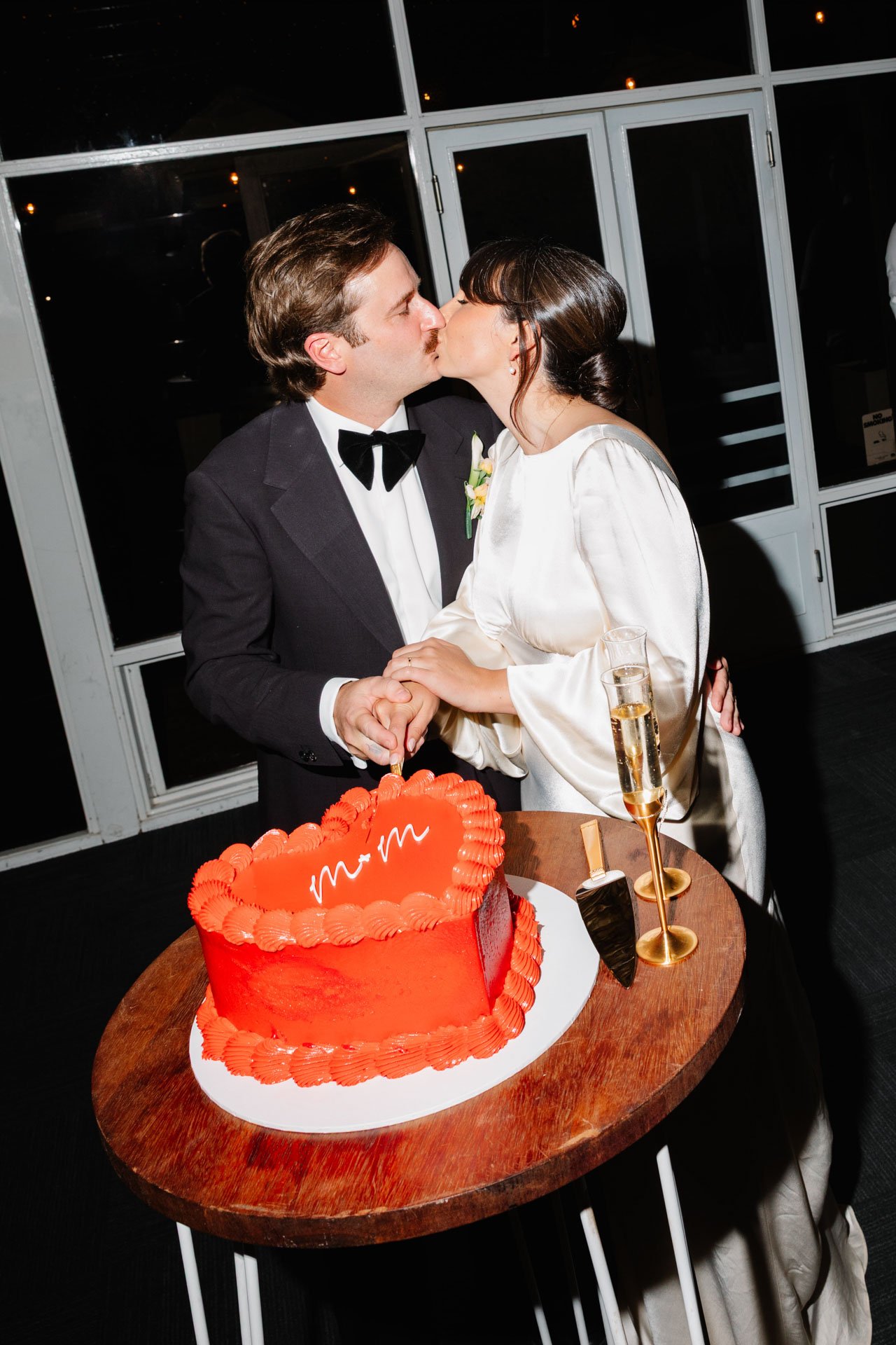 A bride and groom kissing at their wedding reception, holding hands over a round wooden table with a red heart-shaped cake and two champagne glasses.