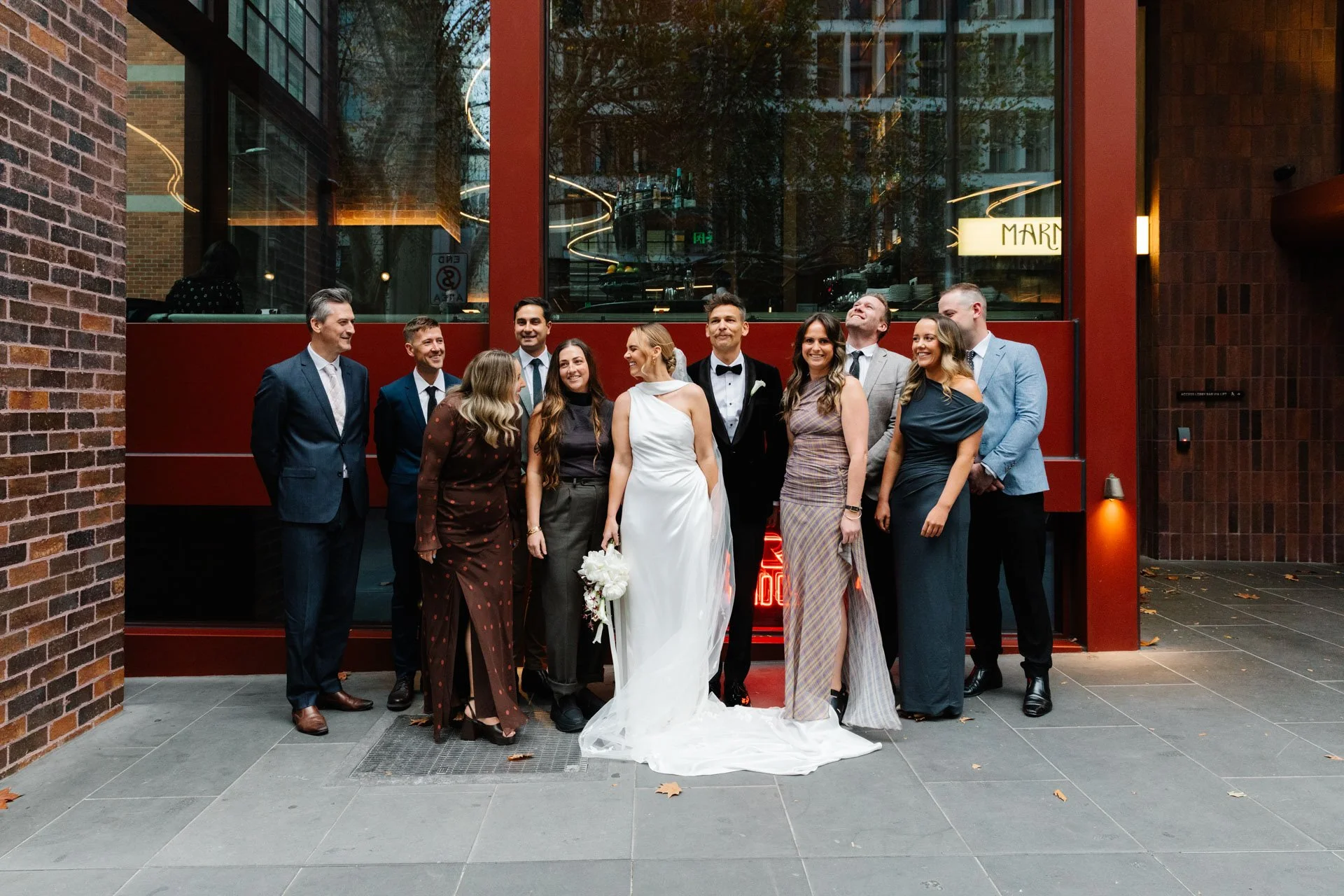 A group of twelve people, including a bride and groom, standing outside a modern building with large glass windows and brick walls. The bride is wearing a white wedding dress and holding a bouquet, and the groom is in a black tuxedo with a bow tie. The group is smiling and dressed in formal attire, celebrating a wedding.