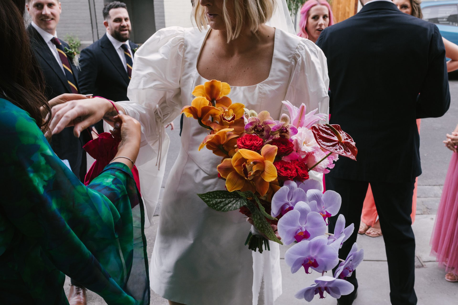 A woman in a white dress holding a large bouquet of colorful flowers at an outdoor event, surrounded by other people dressed in formal attire.