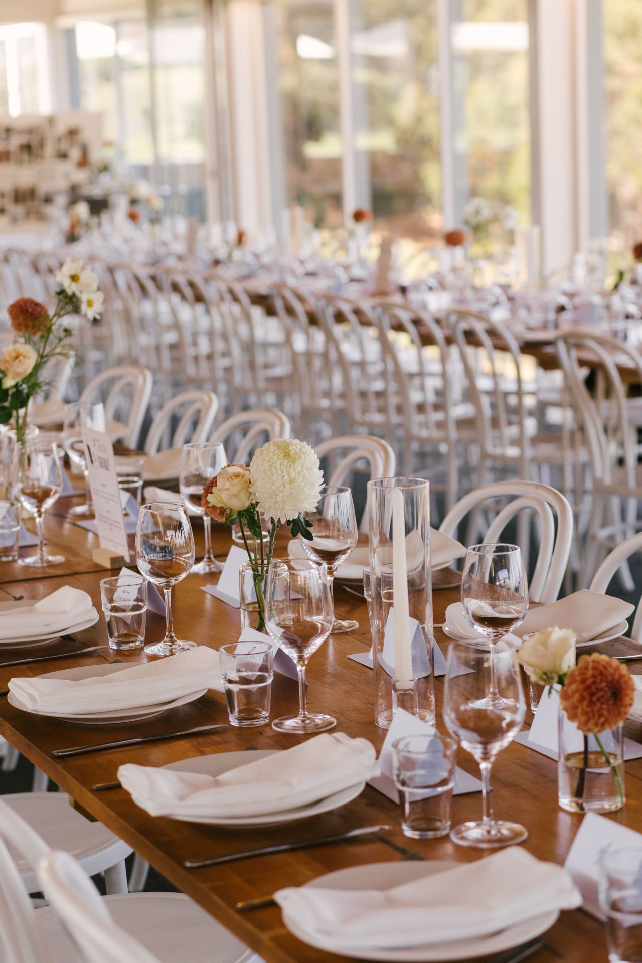 Elegant banquet table set with white plates, silverware, napkins, tall candles, and floral centerpieces in a bright, sunlit room with large windows.