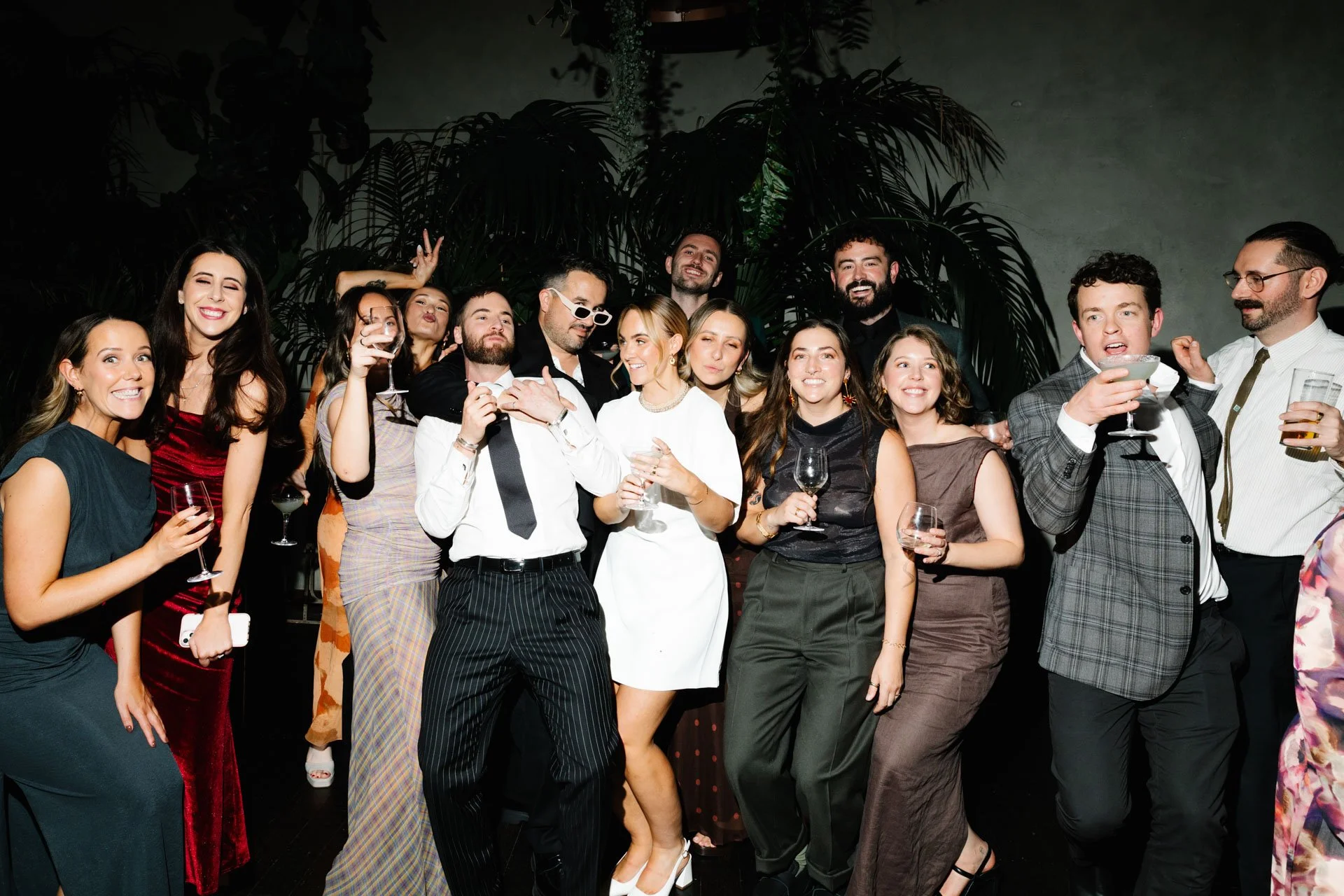 A group of people dressed in party attire enjoying a celebration, holding drinks, smiling, and making gestures in front of a dark leafy background.