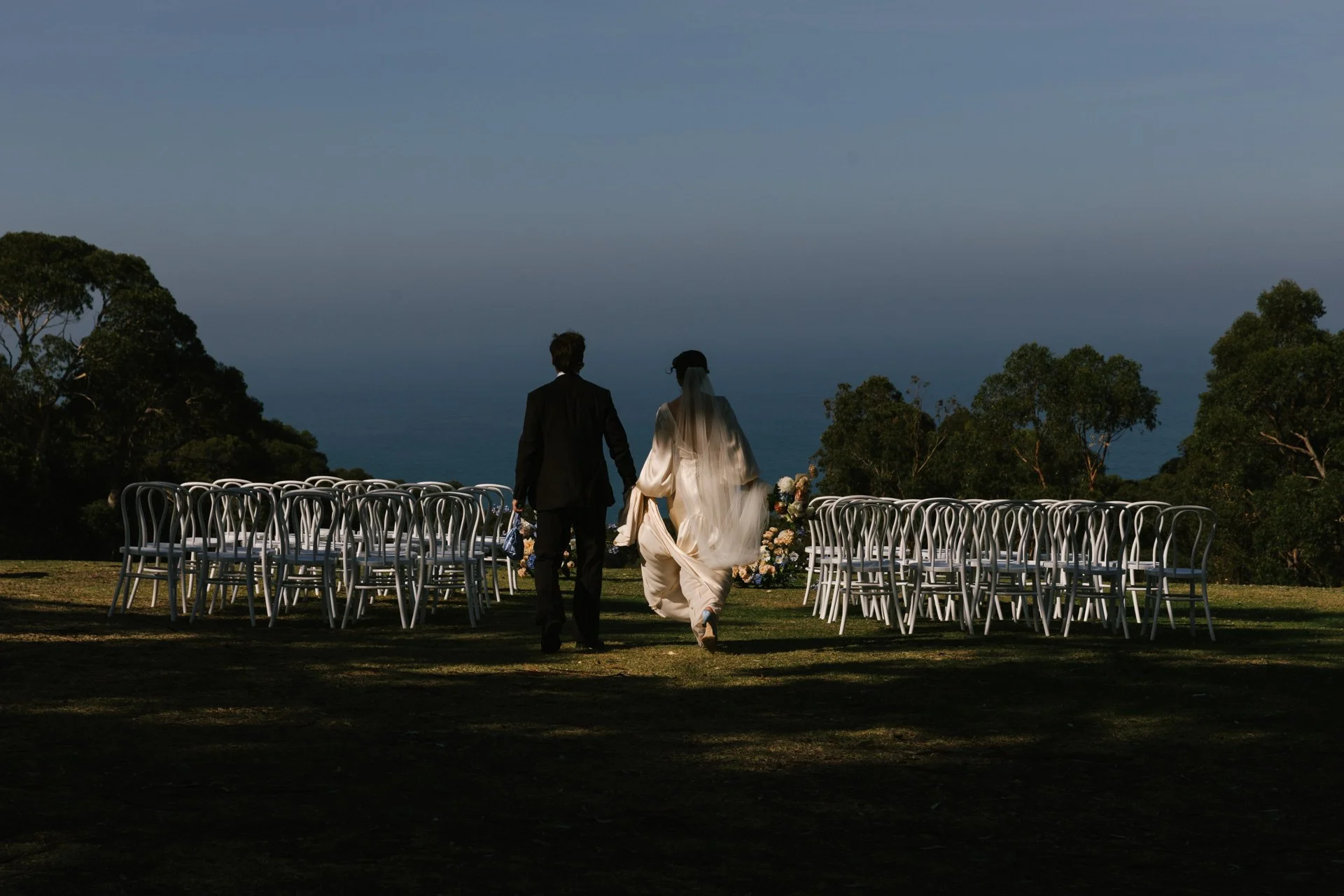 A bride and groom walking hand in hand on a scenic outdoor wedding setup with rows of white chairs and floral arrangements, overlooking a body of water and trees under a cloudy sky.