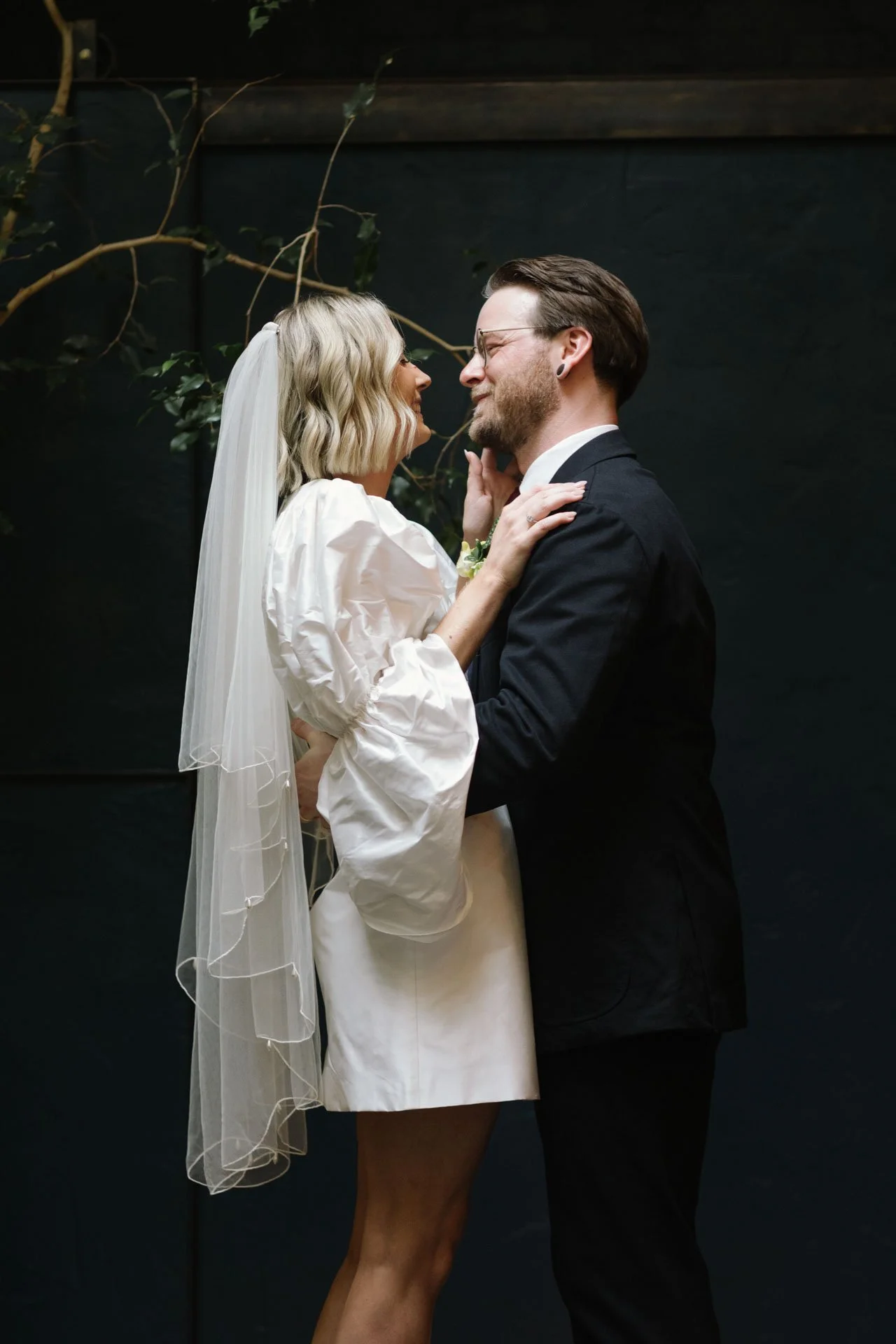 A couple in wedding attire sharing a close moment, with the woman wearing a white dress and veil and the man in a dark suit, against a dark background with plant vines.