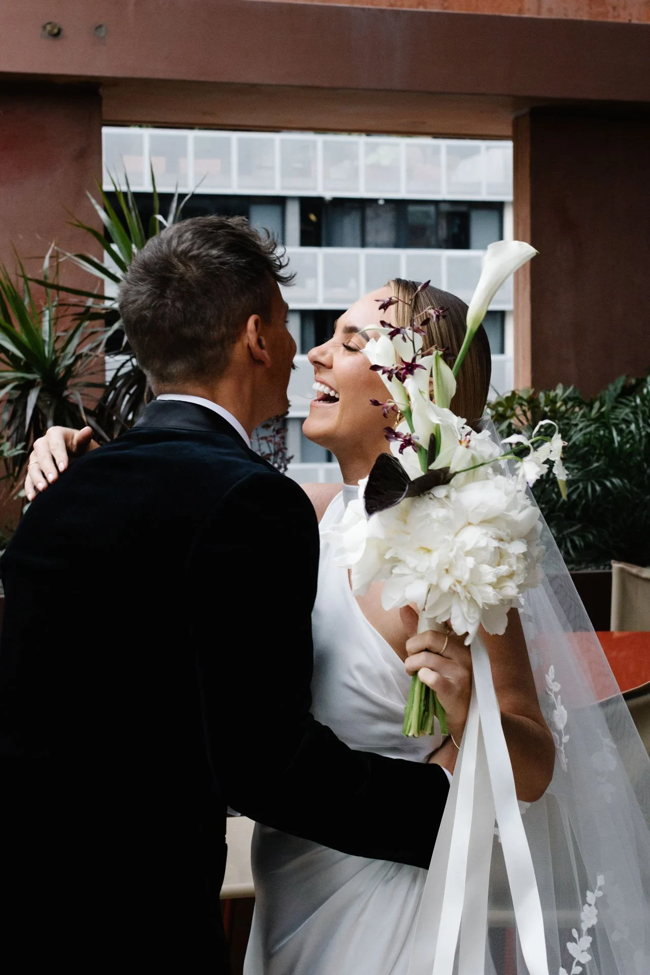 A bride and groom sharing a kiss, with the bride holding a bouquet of white flowers, including calla lilies and orchids, inside a modern building.