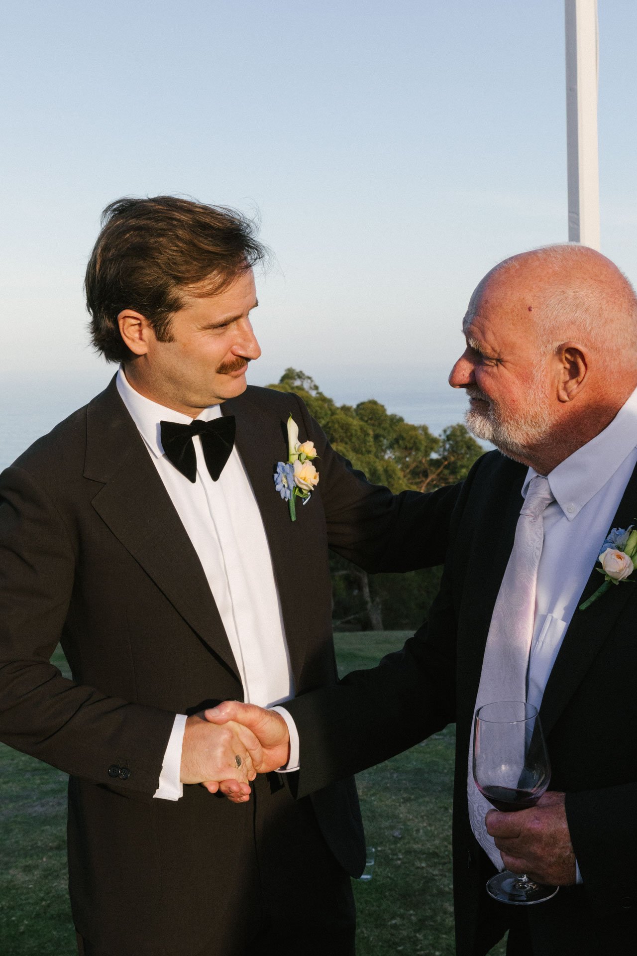 Two men dressed in formal attire, including black tuxedos with white shirts and black bow ties, are shaking hands outdoors during a wedding or celebration. One man has dark hair and a mustache, and the other is bald with a white beard. The man with a