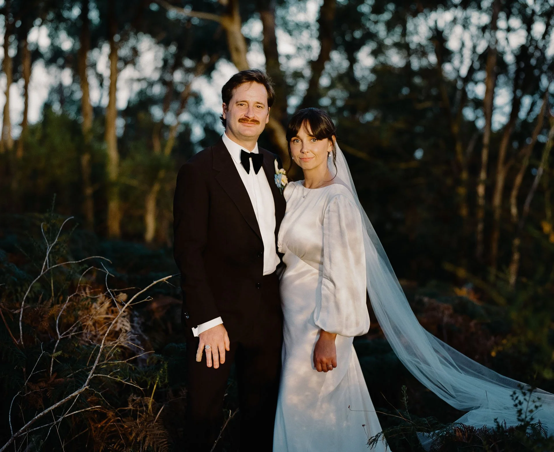 A man and woman dressed in wedding attire standing outdoors in a wooded area during dusk.