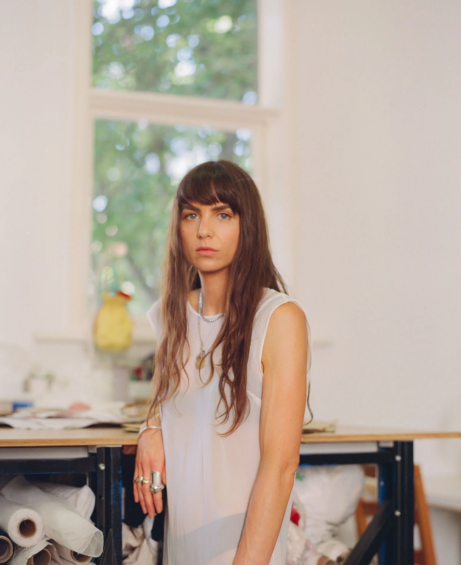A woman with long brown hair and blue eyes in a light-colored sleeveless top standing in front of a window with a blurred outdoor scene. She is indoors, possibly in a creative workspace, with shelves and rolls of paper or fabric visible behind her.