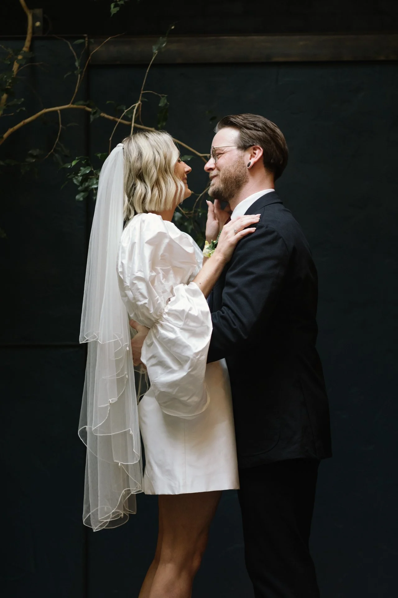 A couple dressed in wedding attire sharing a close, intimate moment against a dark background with some greenery.