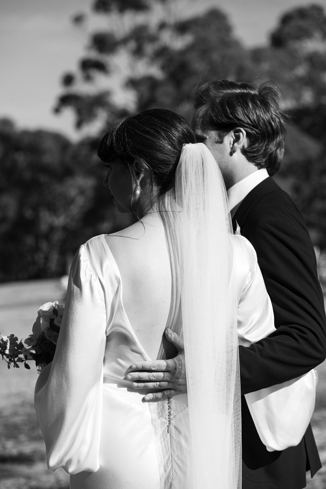 A black and white photo of a bride and groom standing closely together outdoors. The bride wears a wedding dress with a veil, and the groom is in a suit. They are touching foreheads with their eyes closed.