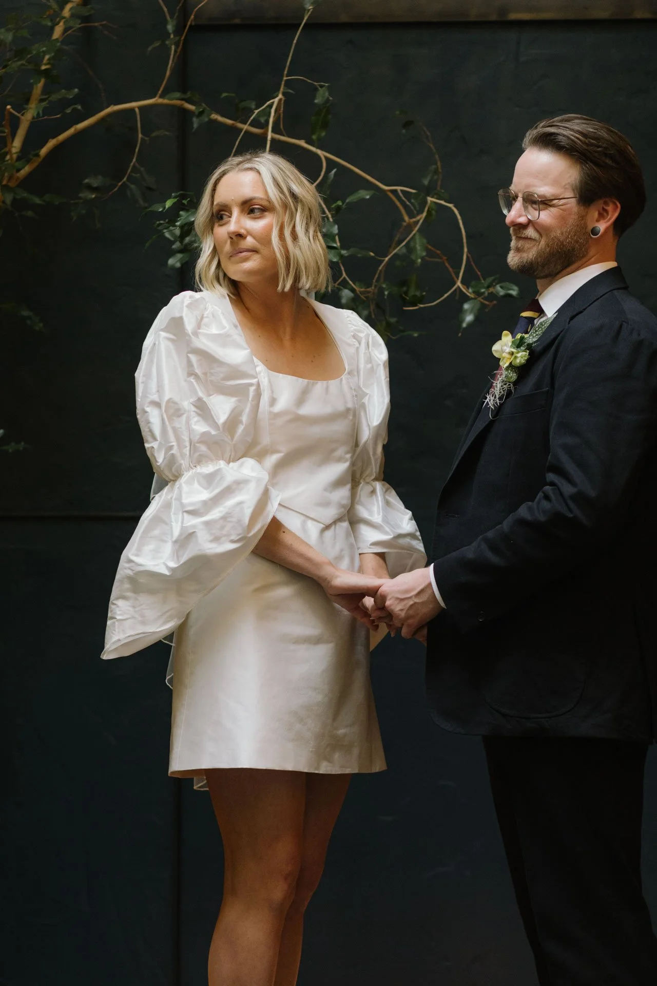 A woman in a white satin dress holding hands with a man in a black suit during a wedding ceremony.