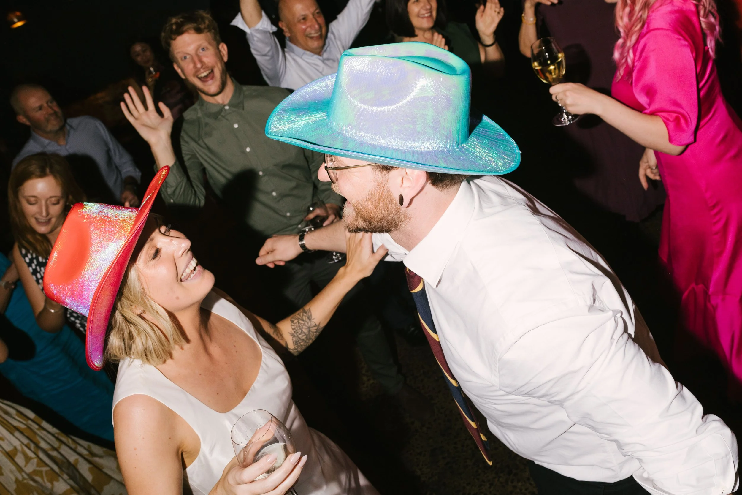 People celebrating at a party, with a woman wearing a pink cowboy hat and a man wearing a large iridescent cowboy hat smiling and dancing.