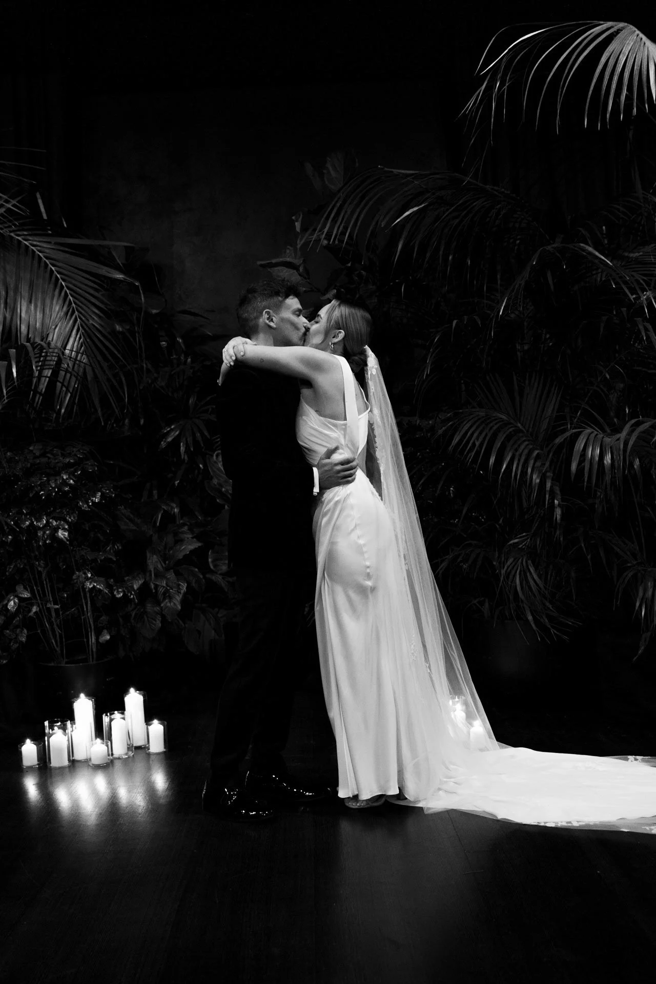 A bride and groom sharing a kiss during their wedding ceremony, with candles on the floor and large leafy plants in the background, in black and white.