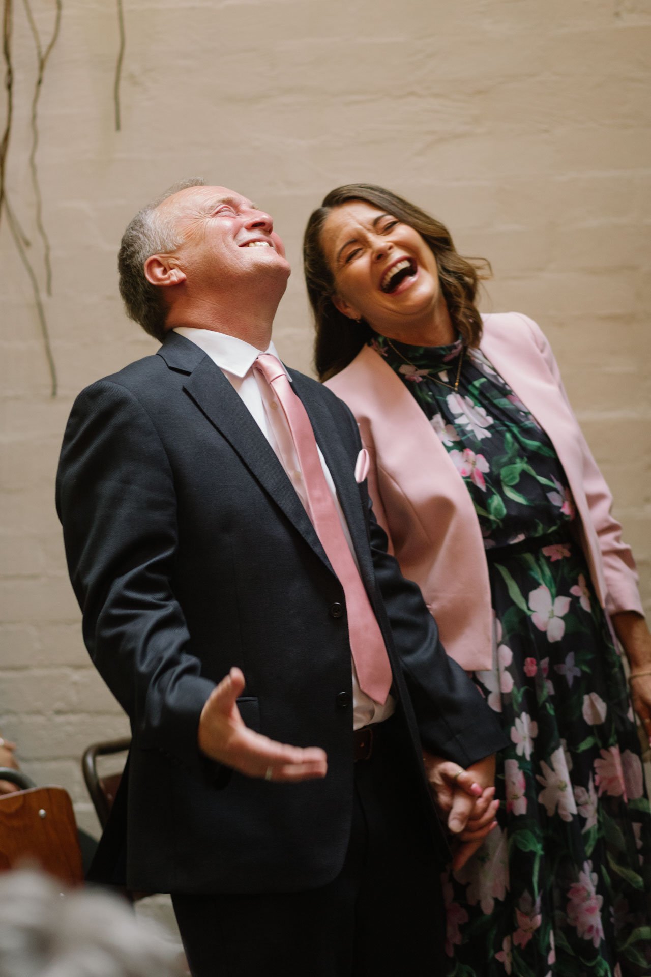 Two people, a man in a suit and a woman in a floral dress with a pink blazer, laughing together in an indoor setting with a beige wall in the background.