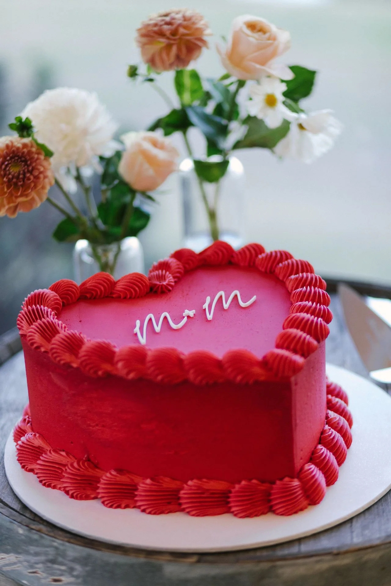 A pink heart-shaped cake with a red piped border and the words 'm + m' written on top, surrounded by a vase of various flowers in the background.
