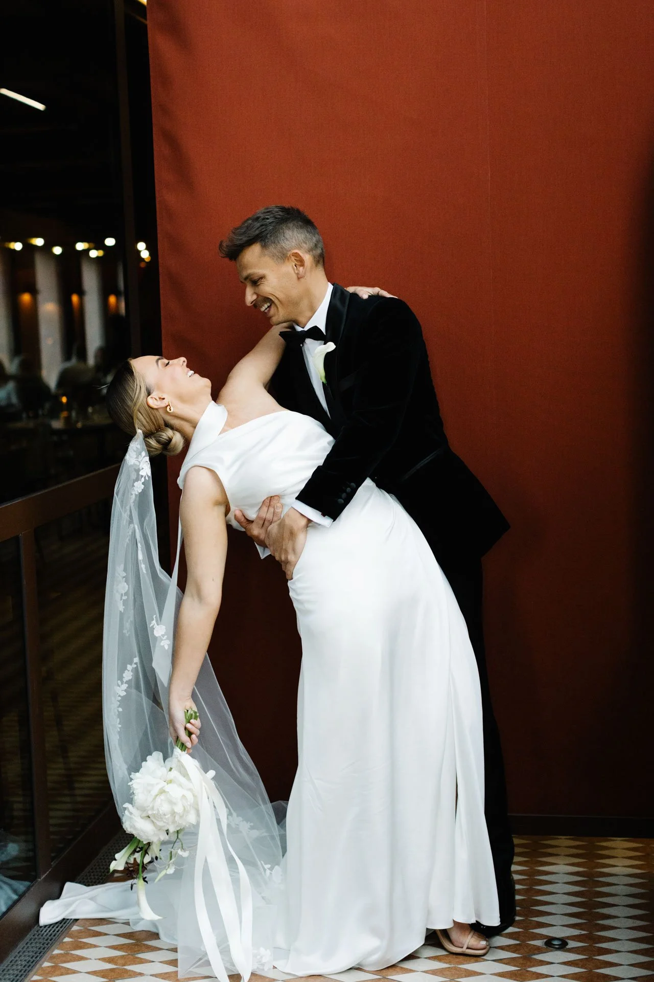 A bride and groom dancing closely at their wedding reception. The bride is in a white wedding dress with a veil, holding a bouquet, and the groom is in a black tuxedo. They are smiling and gazing at each other affectionately.