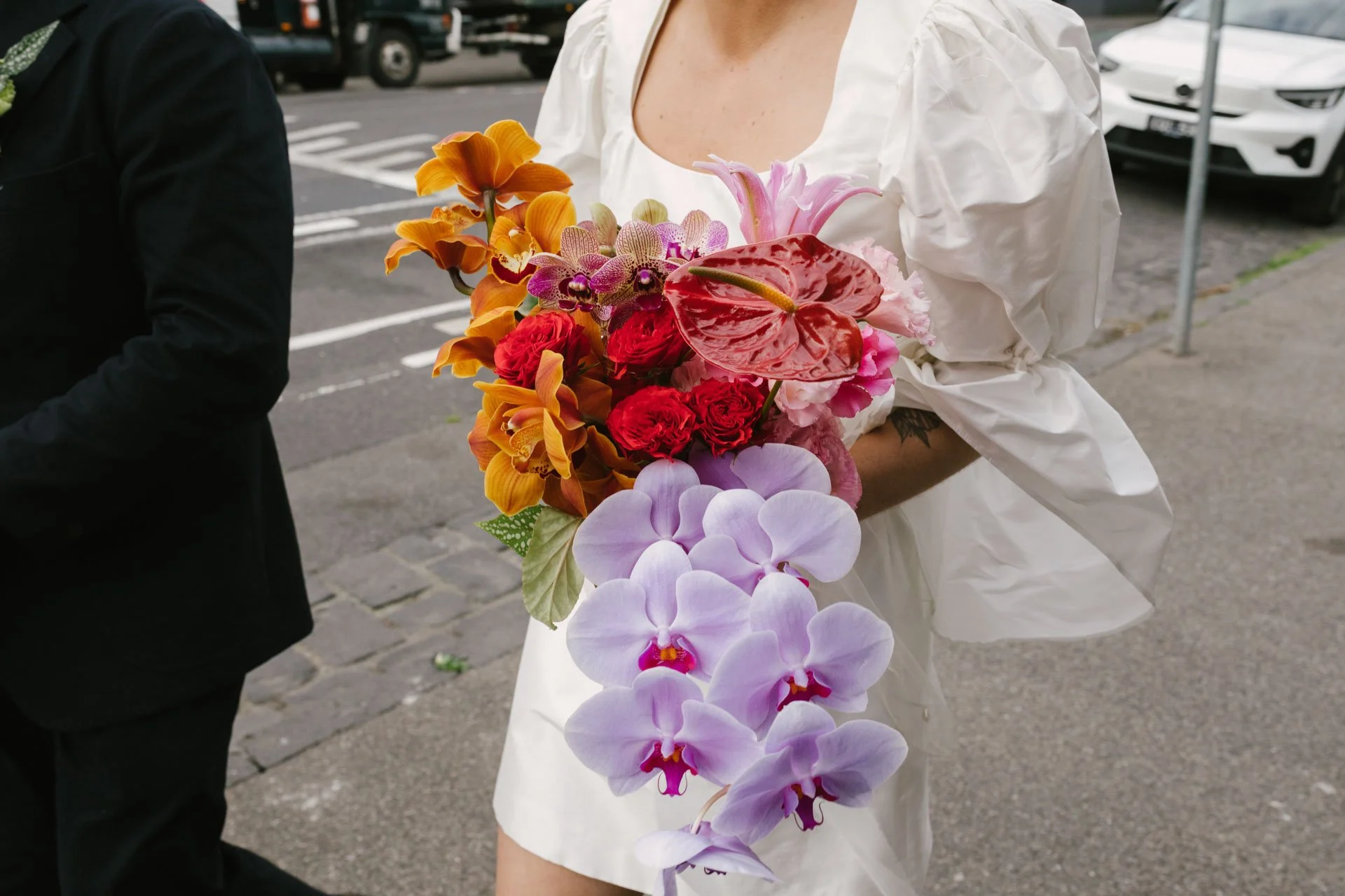 A woman holding a bouquet of colorful flowers including orchids, roses, lilies, and anthuriums, standing outdoors on a city street with parked cars in the background.