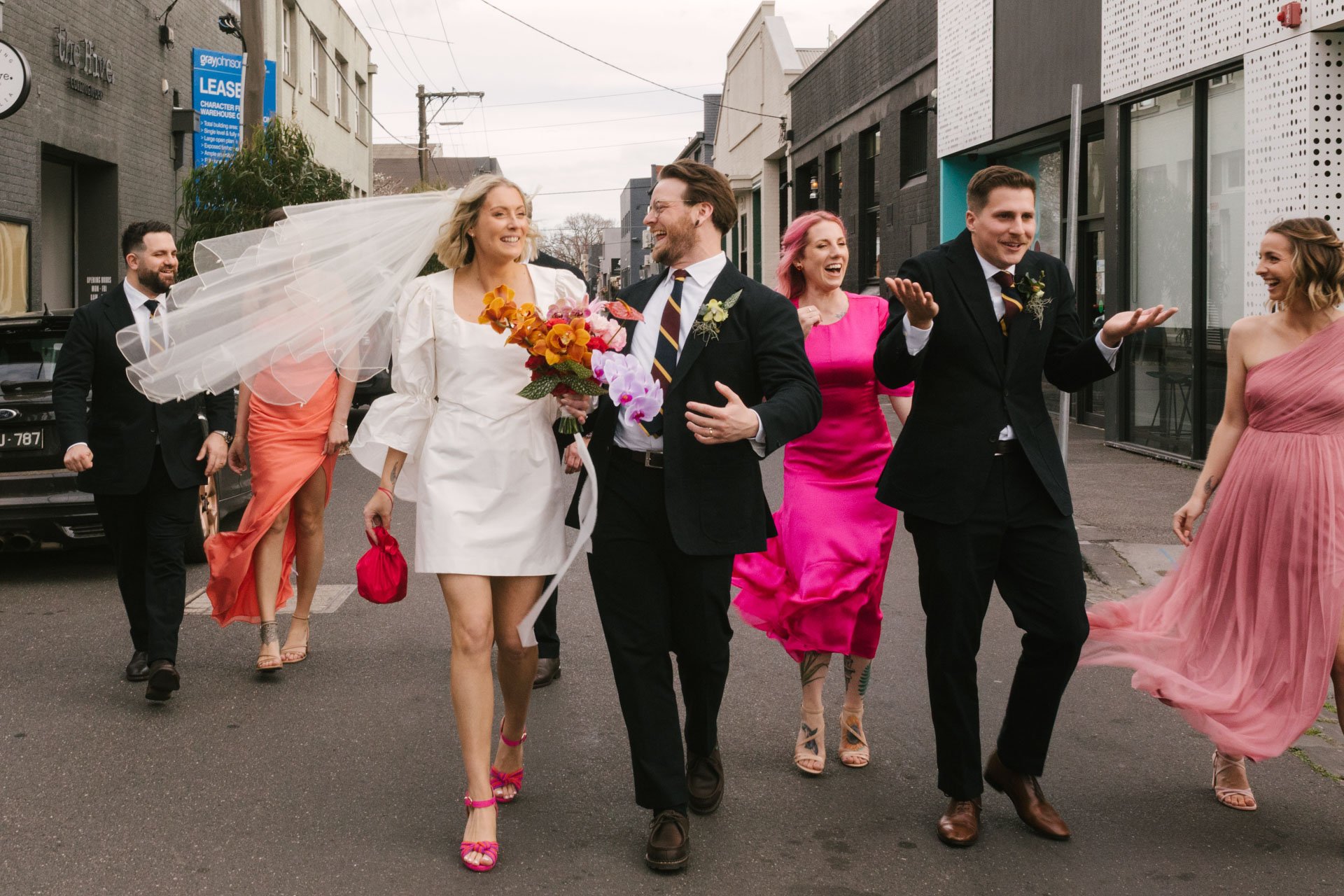 Group of people in formal attire walking together on a city street, celebrating a wedding.