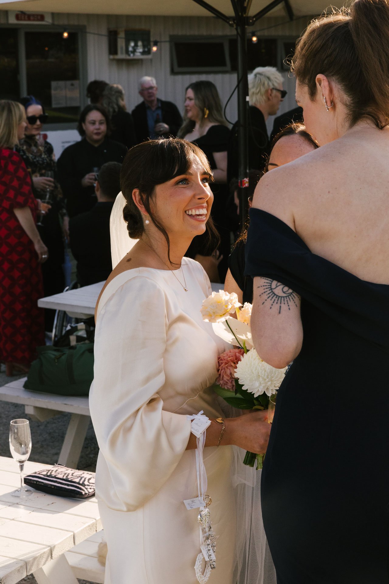 A woman in a white dress holding a bouquet of flowers, smiling and looking at another person during an outdoor event at sunset with a group of people in the background.