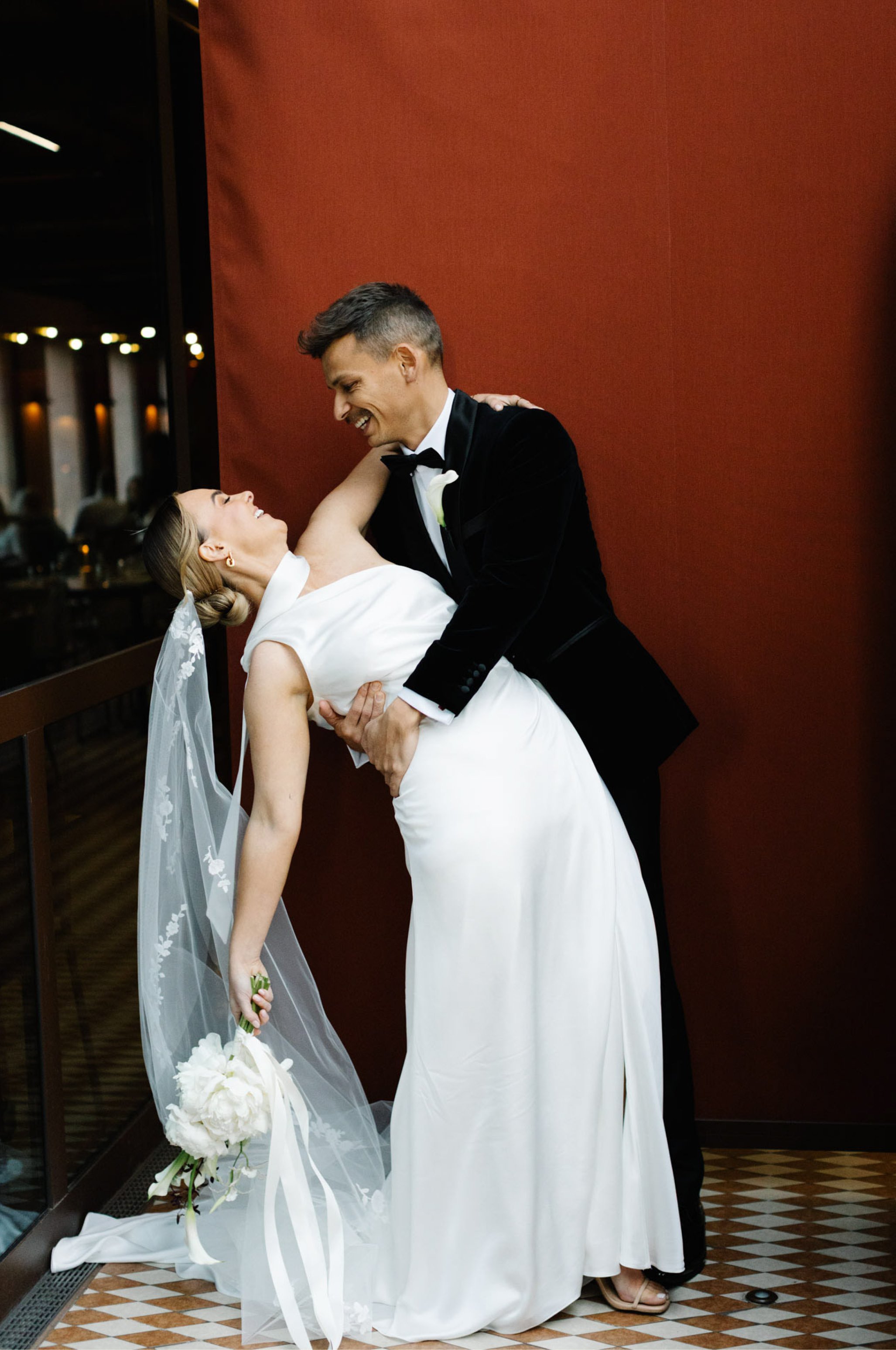A bride and groom dancing at their wedding reception. The groom is holding the bride and smiling, while the bride is laughing. The bride is wearing a white dress and veil, holding a bouquet of white flowers. They are standing on a checkered floor against a red wall.