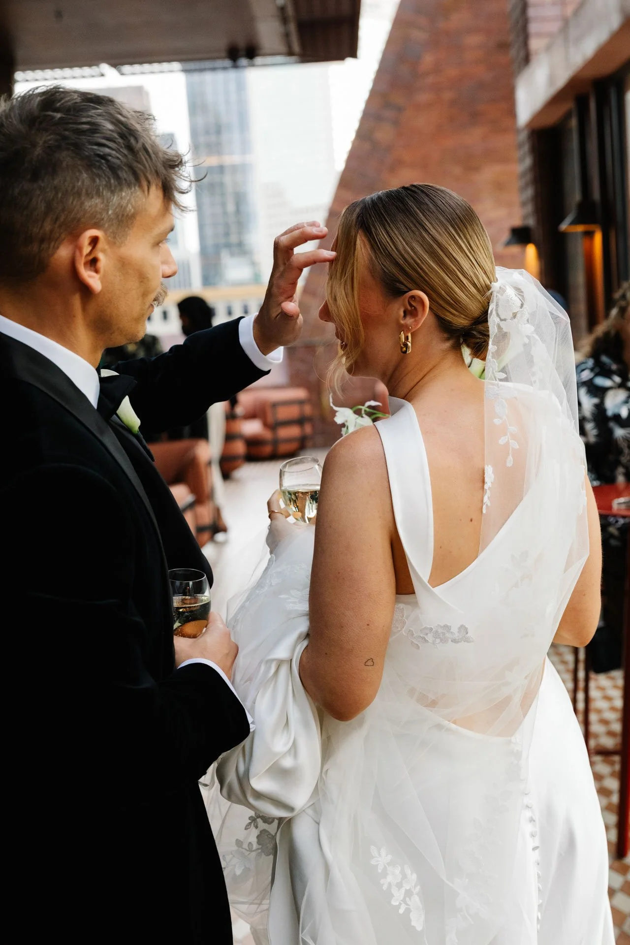 A bride and groom at their wedding, with the groom gently touching the bride's forehead while holding a glass of champagne, in an urban venue with brick walls and large windows.