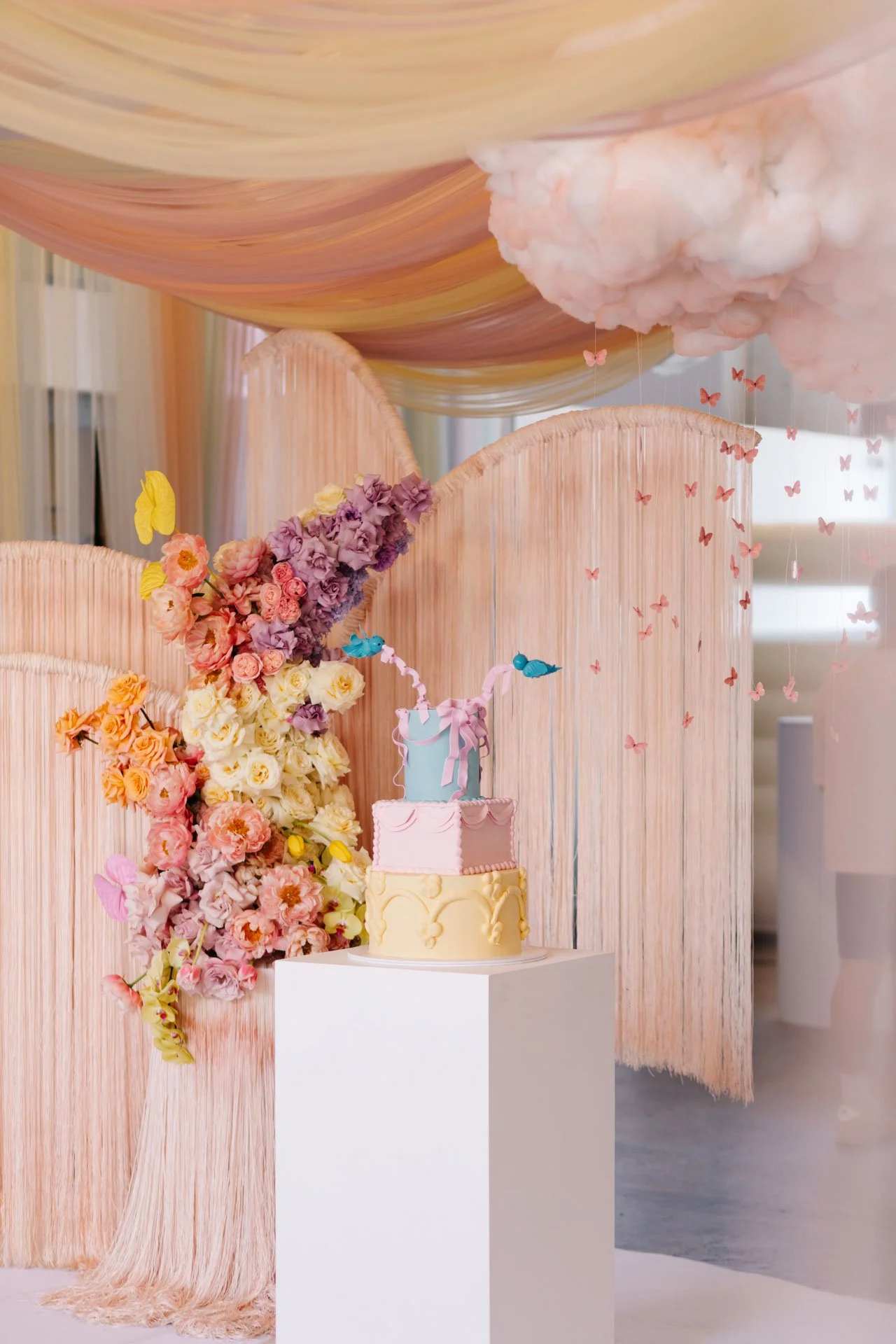A pastel-colored birthday cake decorated with a unicorn horn, ears, and flowers, displayed on a white pedestal against a backdrop of pink and peach drapes and floral arrangements, with pink butterfly decorations hanging.
