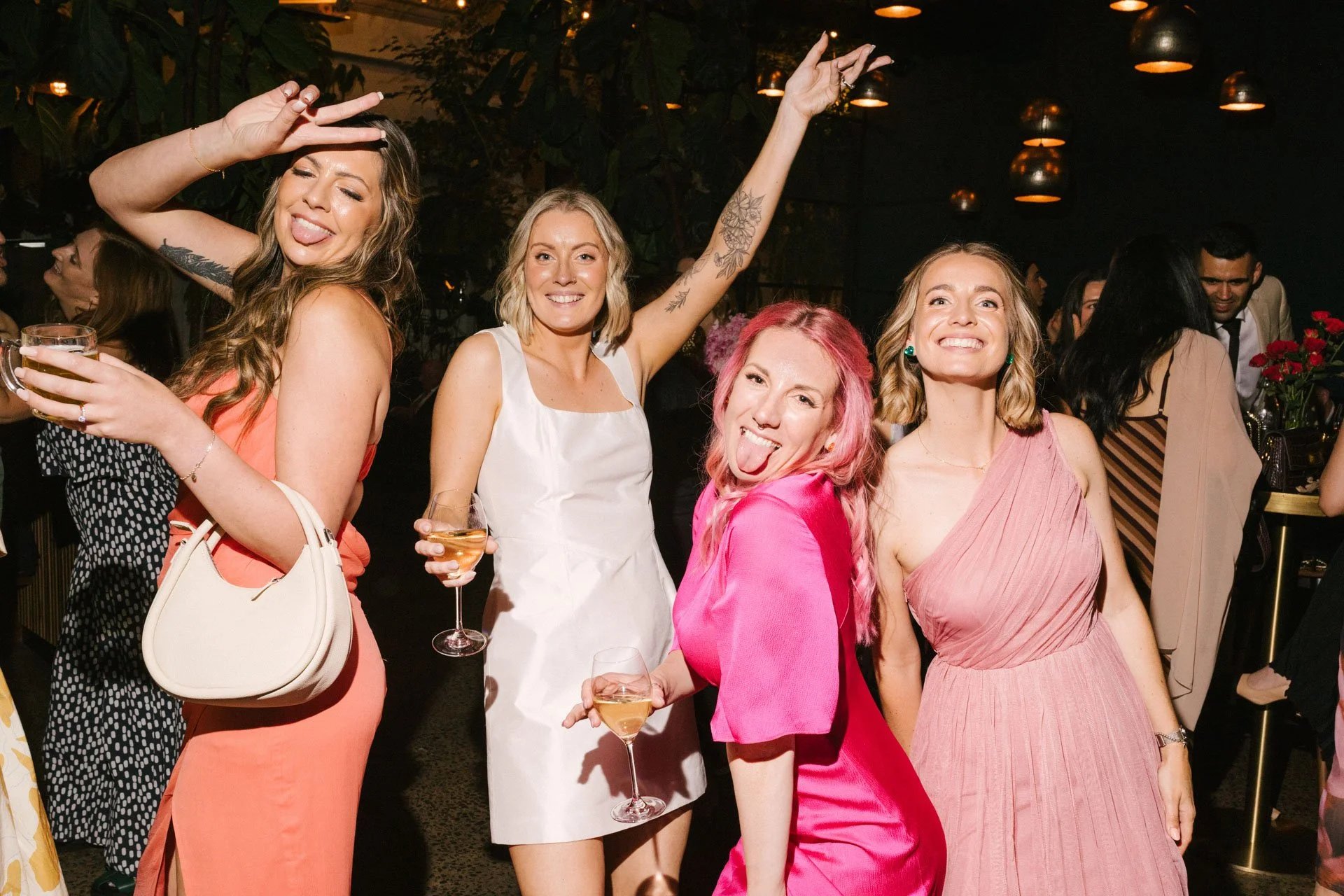 A group of women celebrating at a party, smiling, holding drinks, and making playful gestures. The background shows a lively indoor setting with other people and decorative lighting.