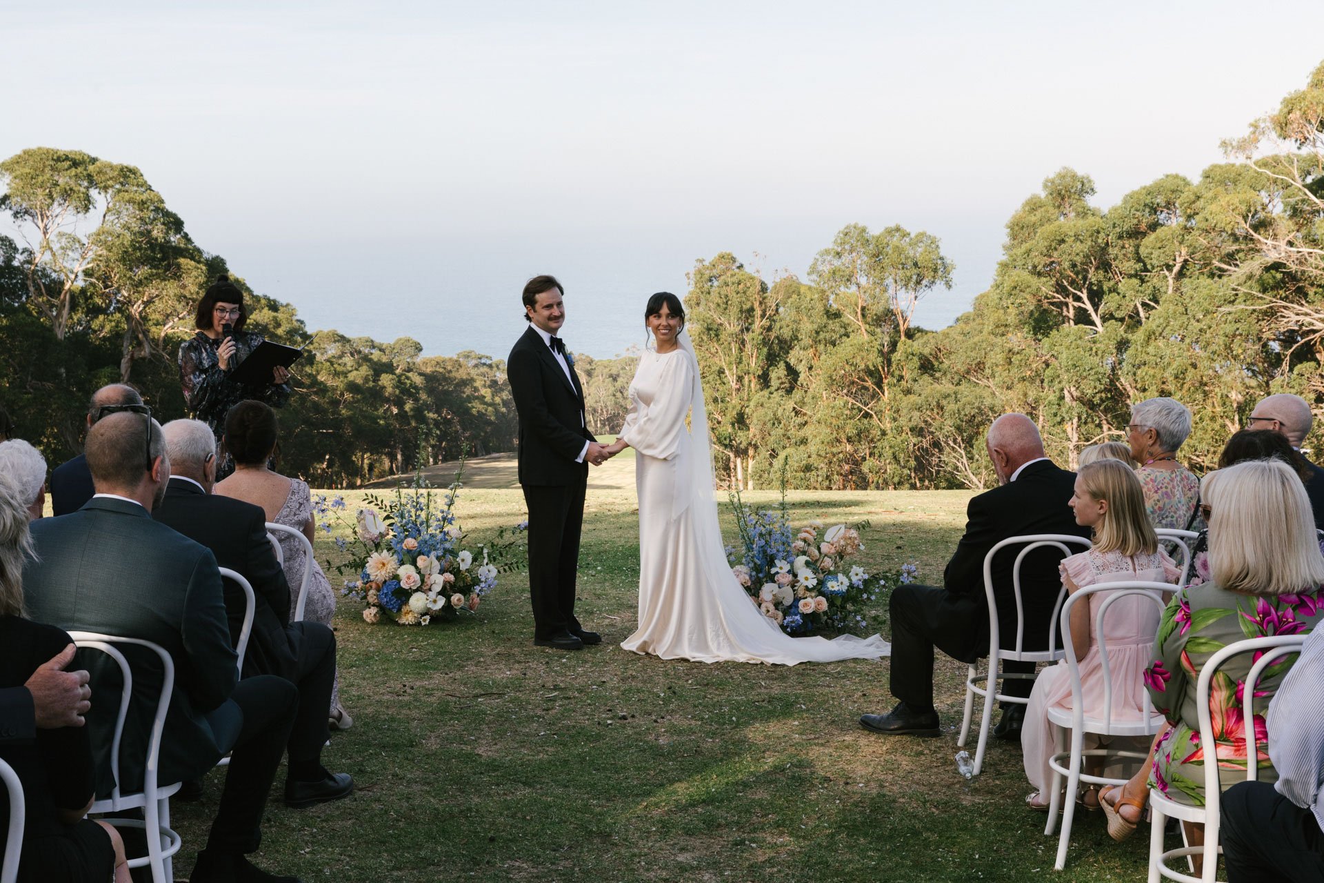 A wedding ceremony taking place outdoors on grass, with a bride and groom holding hands at the altar, surrounded by seated guests and a floral arrangement. There are trees and an open sky in the background.