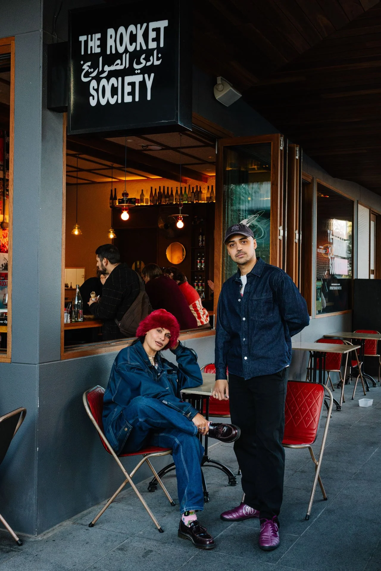 Two fashionably dressed individuals, one seated with red hair and casual attire including a blue jacket and jeans, and the other standing wearing a dark denim jacket, black pants, and purple sneakers, pose outside a cafe named 'The Rocket Society' wi