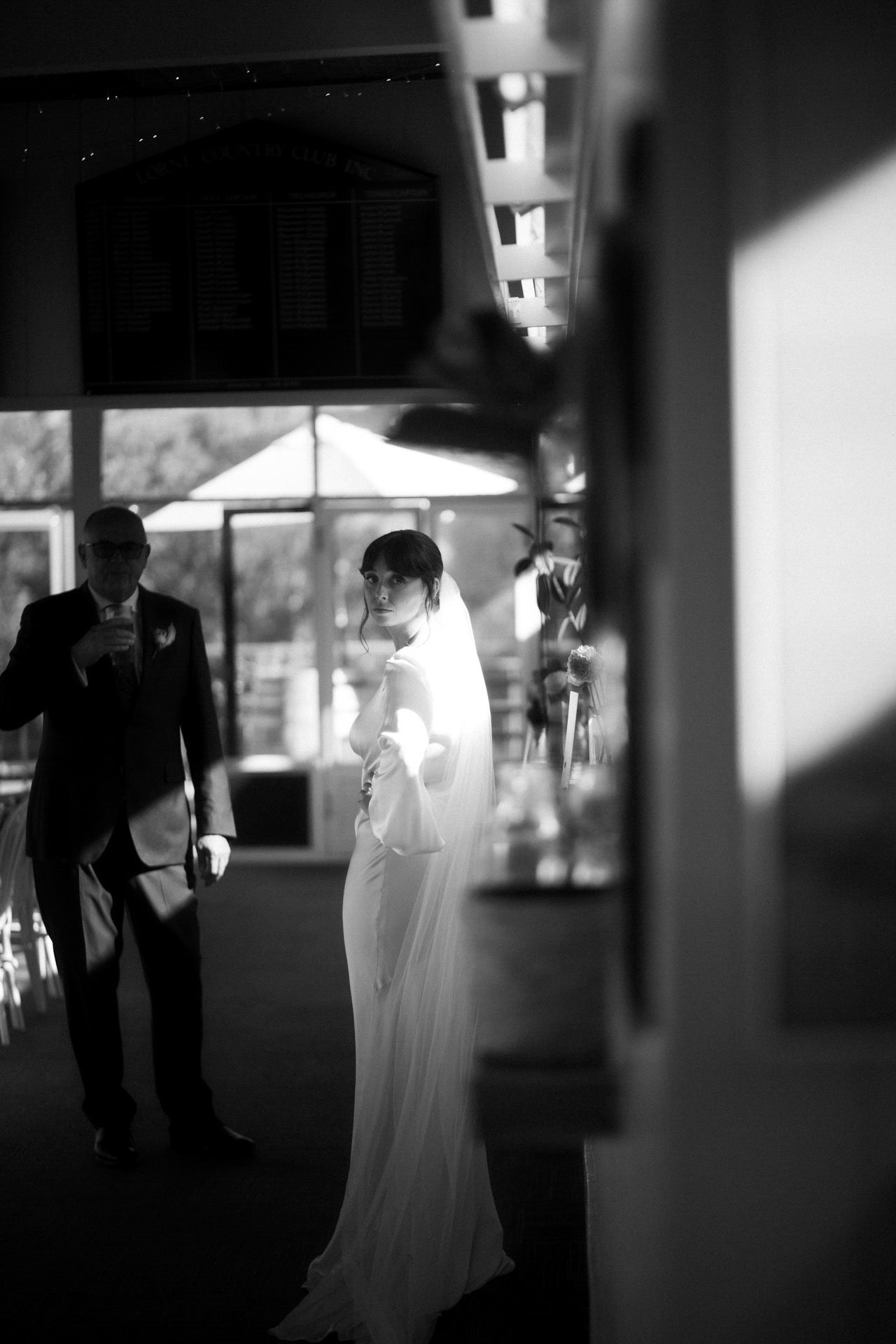 Black and white photo of a bride in a wedding gown standing indoors, looking towards the camera, with a man in a suit behind her holding a drink, possibly at a wedding reception.